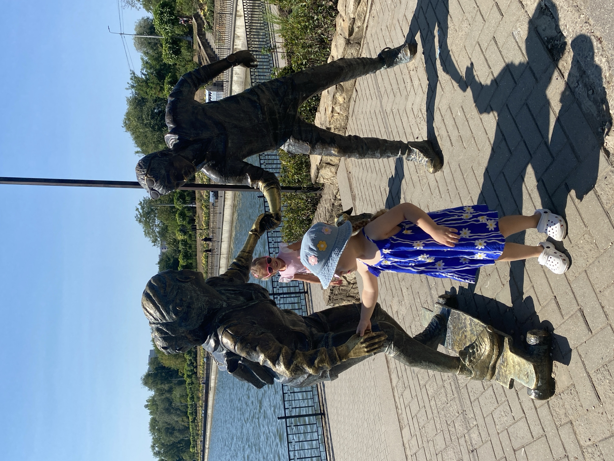 Two children, a young girl wearing a blue floral dress and a bucket hat, and an older girl with pink glasses and a pink top, interact with a bronze sculpture of two individuals on a skateboard. The sculpture depicts a male and female figure. The girl in the blue dress is reaching out to touch the sculpture. The older girl stands behind the sculpture. They are located near a body of water with a fence and trees in the background. This sculpture is located in the Valea Morilor Park in Chișinău, Moldova.