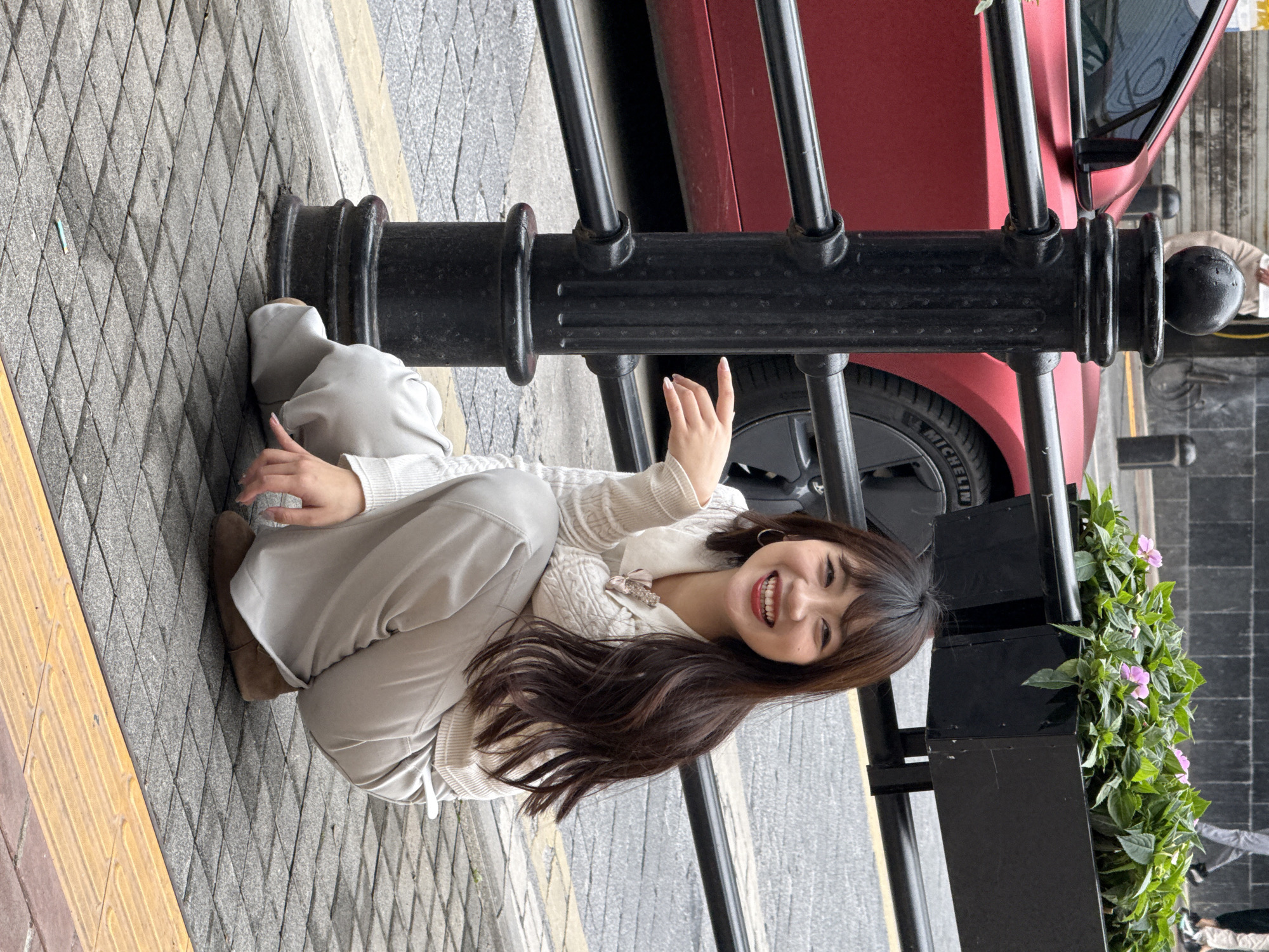 A woman is sitting on the ground next to a black metal fence and a red car in Yuzhong District, China. The ground is paved with gray and yellow stones. The woman is wearing a white sweater and light gray pants. She is smiling and waving. A planter box with greenery and pink flowers is visible to the right of the fence. The background suggests an urban environment.