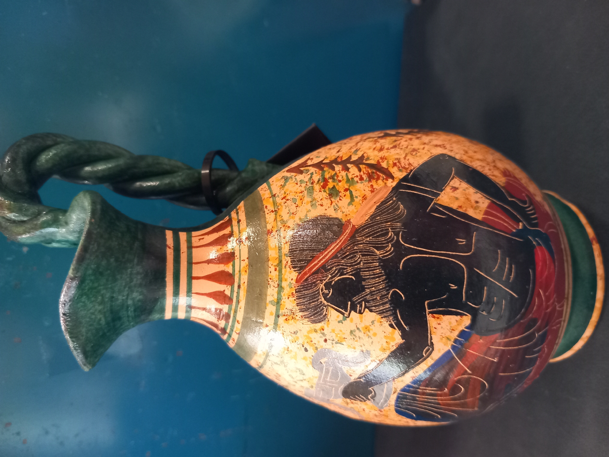 This is a close-up, eye-level shot of a decorative amphora, likely a replica, placed on a dark surface. The amphora has a light-colored, speckled body adorned with a black silhouette of a seated figure with a long beard and hair, wearing a red headband. The figure is holding a light blue object. The amphora also features red and blue painted drapery below the figure, and decorative bands of green, orange, and red around its neck and base. The handle of the amphora is a thick, twisted green rope-like shape. The background is a deep blue, textured wall. The lighting is bright, creating reflections on the glossy surface of the amphora.