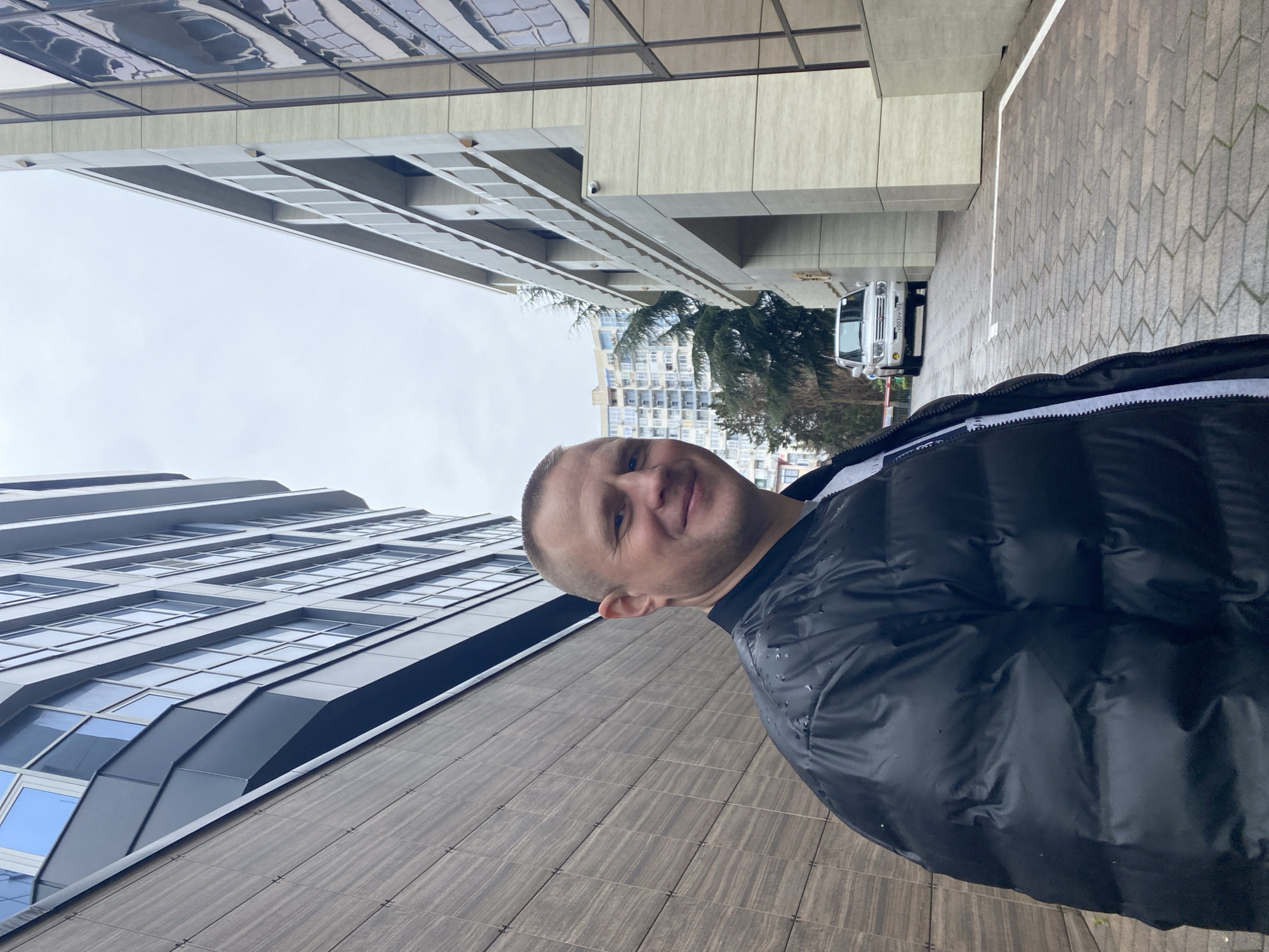 A man stands in an alleyway between two high-rise buildings, a dark puffer jacket on his person. The building to his left is grey, modern, with rows of tinted windows. The building to his right is a lighter beige color, with a window grid pattern. The alleyway has grey tiled ground. A white SUV is parked further down the alleyway. The man is smiling.
The image was taken in the city of Sochi, Russia.