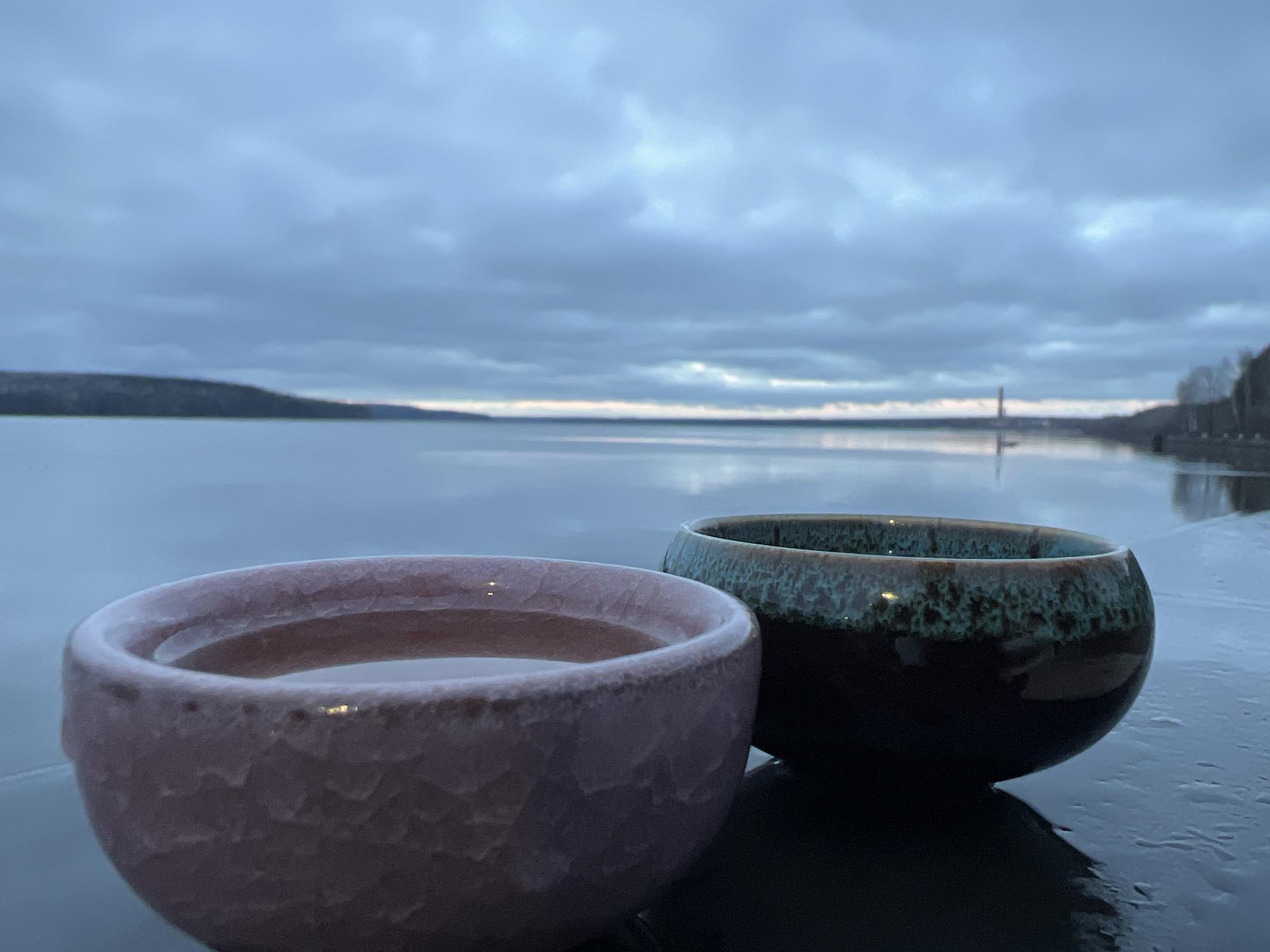 The image shows two cups on a wet, reflective surface, with a blurred background of a body of water, a distant shoreline with trees, and a cloudy sky. The cup in the foreground on the left is light pink, and filled with a clear liquid. The cup to the right has a dark brown bottom and a teal-colored rim. In the background, the body of water stretches toward the horizon, and there's a structure resembling a tower in the distance. The location is Chaikovsky, Russia.