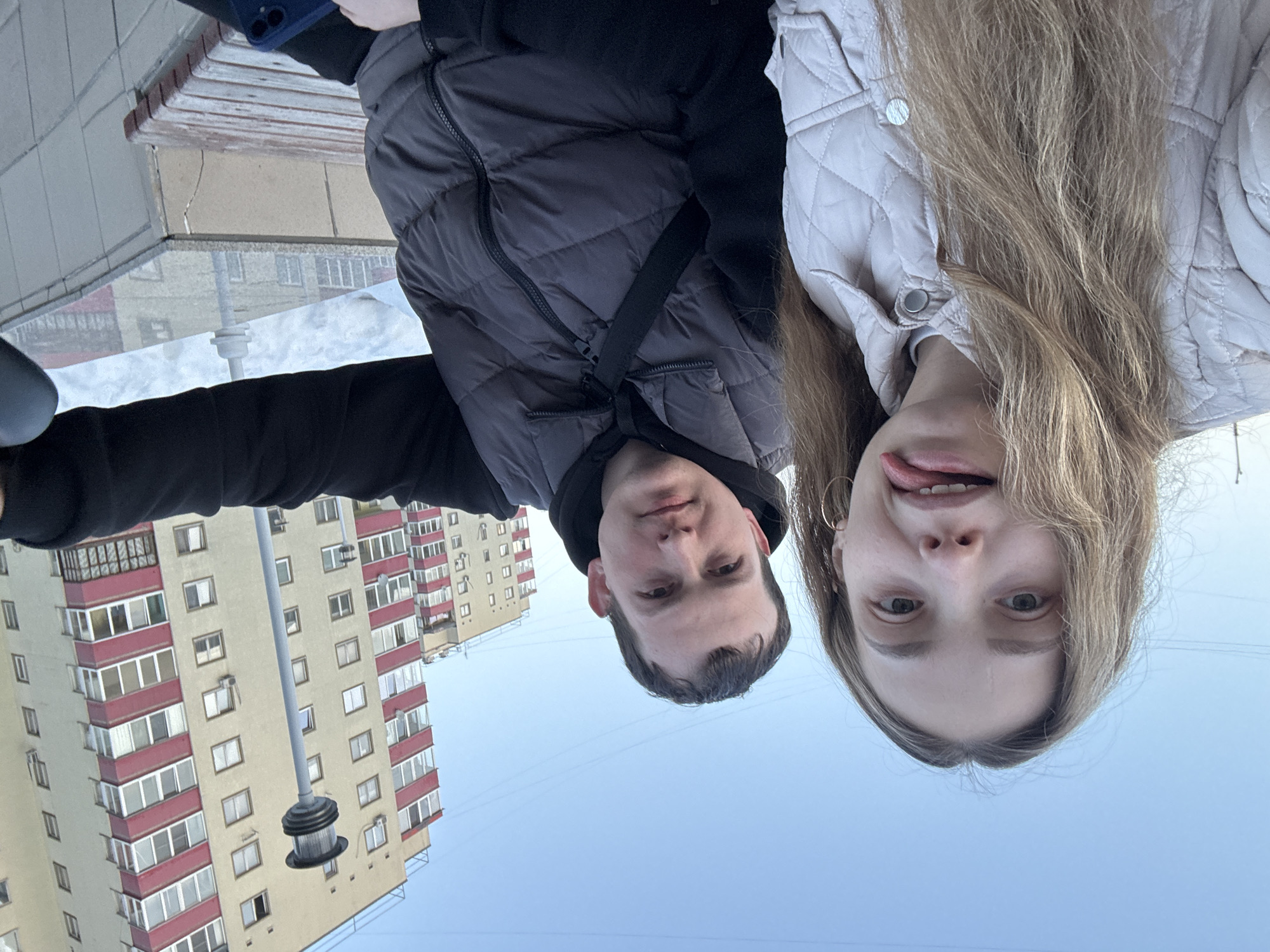 Two individuals are seated outdoors in Chaikovsky, Russia, facing the camera. A female on the left has long, light brown hair, wears a light-colored quilted jacket with silver buttons, and gold hoop earrings. She is looking directly at the camera with her tongue extended. A male on the right, with short dark hair, wears a dark grey puffer vest over a black hooded sweatshirt, with a black strap crossing his chest. He also looks directly at the camera, maintaining a neutral expression. A dark blue smartphone is visible in his left hand, resting on his lap.
Both subjects are positioned in front of a partially visible wooden slatted bench. The background features a tall, multi-story residential building with cream-colored walls and prominent red balcony sections, set against a clear blue sky. A grey lamppost with a multi-tiered circular fixture stands in front of this building. A second, similar residential building is partially visible further back on the left. A large pile of snow is present in the middle ground to the right. Overhead power lines are faintly visible against the sky.