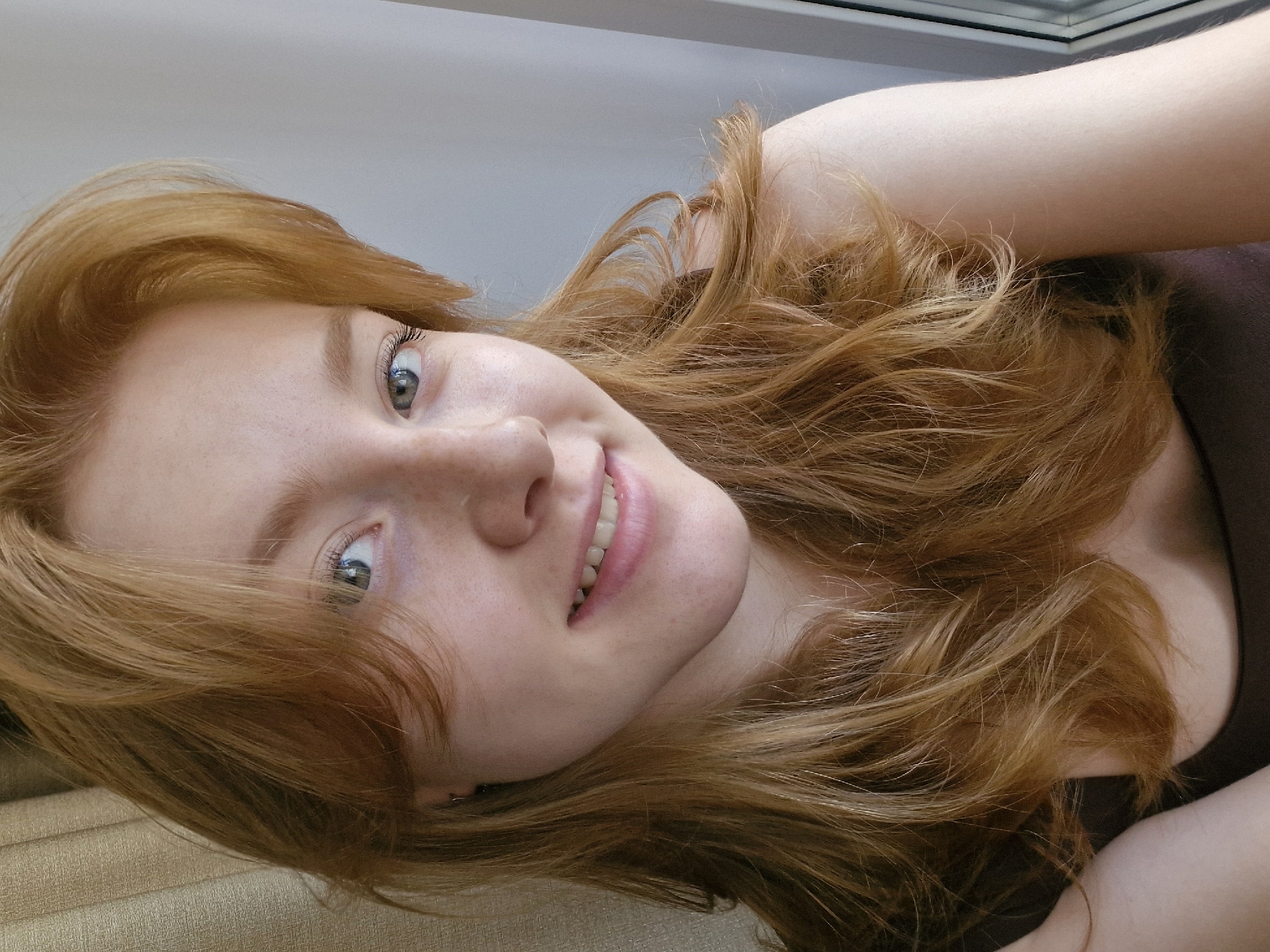 A close-up portrait of a young woman with reddish-blonde hair. The woman is smiling, her head tilted slightly. Freckles are visible on her face. She is wearing a dark-colored top. The background appears to be a light-colored wall. The location is unknown.