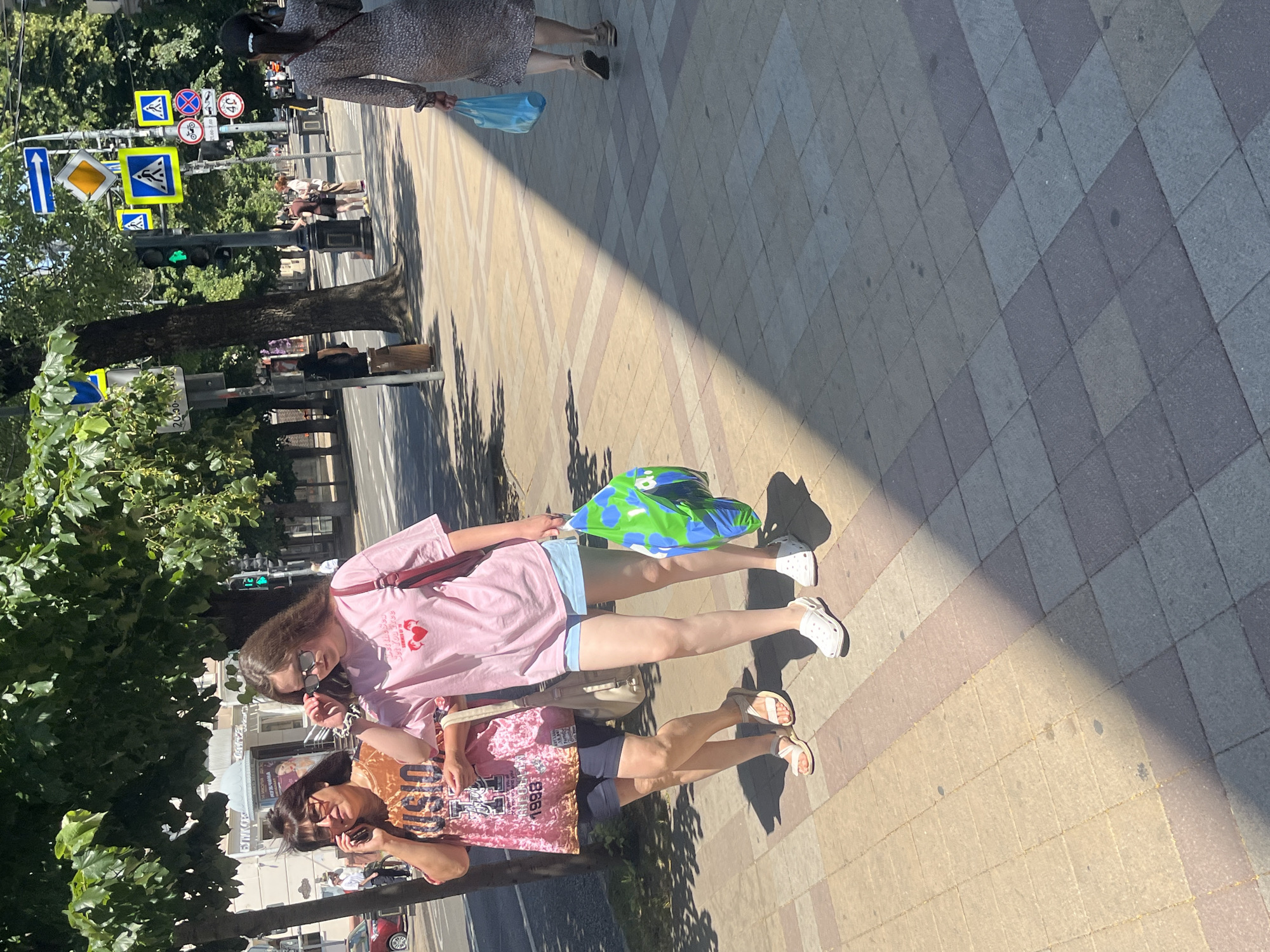 Two women are walking on a paved sidewalk in Krasnodar, Russia. The woman on the left is wearing a pink graphic t-shirt and dark shorts, talking on her phone. She has dark hair and is wearing glasses and sandals. The woman on the right is wearing a pink t-shirt, blue shorts, and white clogs. She has long brown hair and is carrying a multi-colored shopping bag. Behind them, a woman in a patterned dress is walking away, carrying a blue plastic bag. The scene is on a sunny day, with trees and buildings visible in the background. Traffic lights and street signs are also present, indicating an urban environment.