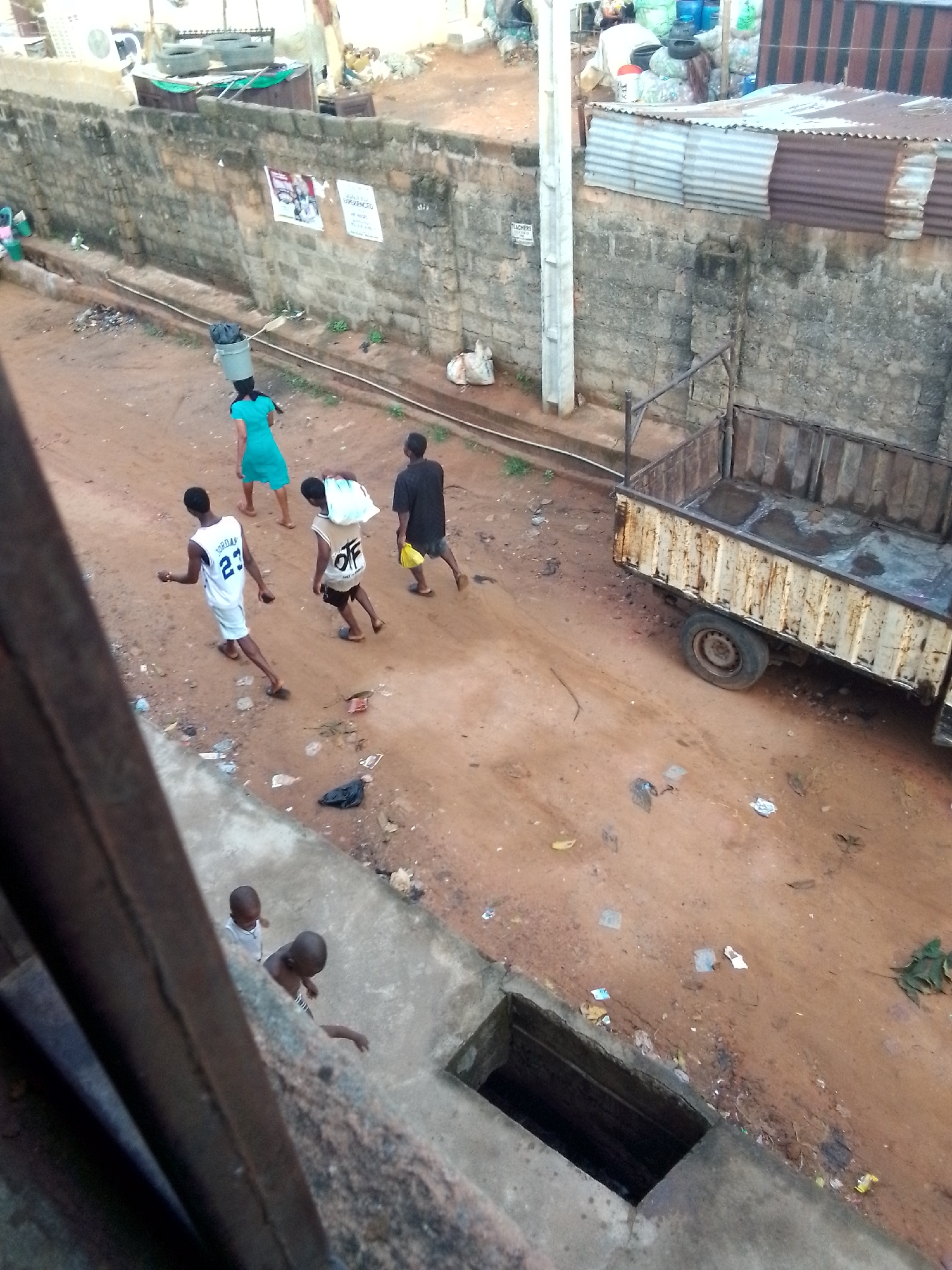 The image shows an outdoor scene in what appears to be a neighborhood in Unknown, Unknown. There are four individuals walking down a dirt road. One is wearing a teal dress and carrying a bucket on her head. Two males are walking together, one in a white jersey with the number 23 on it and the other carrying a white bag over his shoulder. The last individual is walking behind them in a dark-colored shirt. To the right of the road is a rusted, flatbed trailer. In the foreground, near the bottom of the frame, two younger individuals are looking over a concrete structure with a square opening, possibly a drain or access point. The area is marked by litter and debris. A tall brick wall runs along one side of the road. Overhead, the perspective suggests the photo was taken from a higher vantage point, potentially from a building's upper floor.