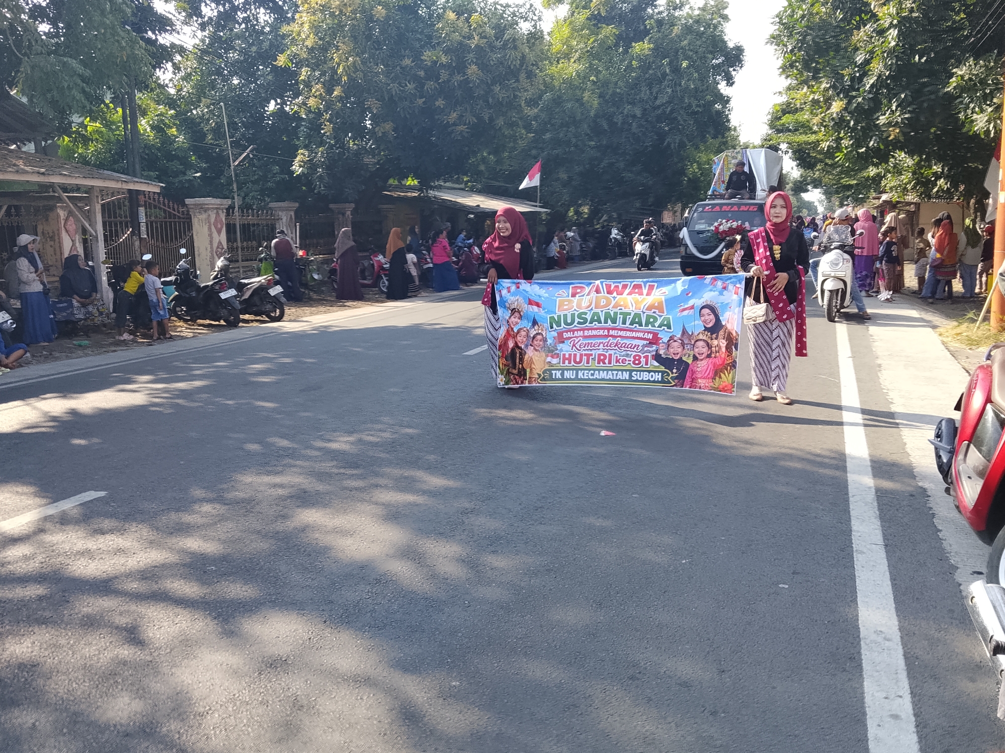 In Buduan, Indonesia, on a sunlit day, a parade is underway on an asphalt road lined with trees and buildings. A banner reading "PAWAL BUDAYA NUSANTARA DALAM RANGKA MEMERIAHKAN KEMERDEKAAN HUT RI KE-81 TK NU KECAMATAN SUBOH" is held by a woman in a maroon headscarf and a black top with a decorative sash. Behind her, a truck with a banner that reads "LANANG" is visible, adorned with red and white flowers. Numerous people are gathered on the side of the road, some observing the parade, while others are on motorcycles and scooters. Children in colorful clothing are also present. The environment suggests a festive community event.