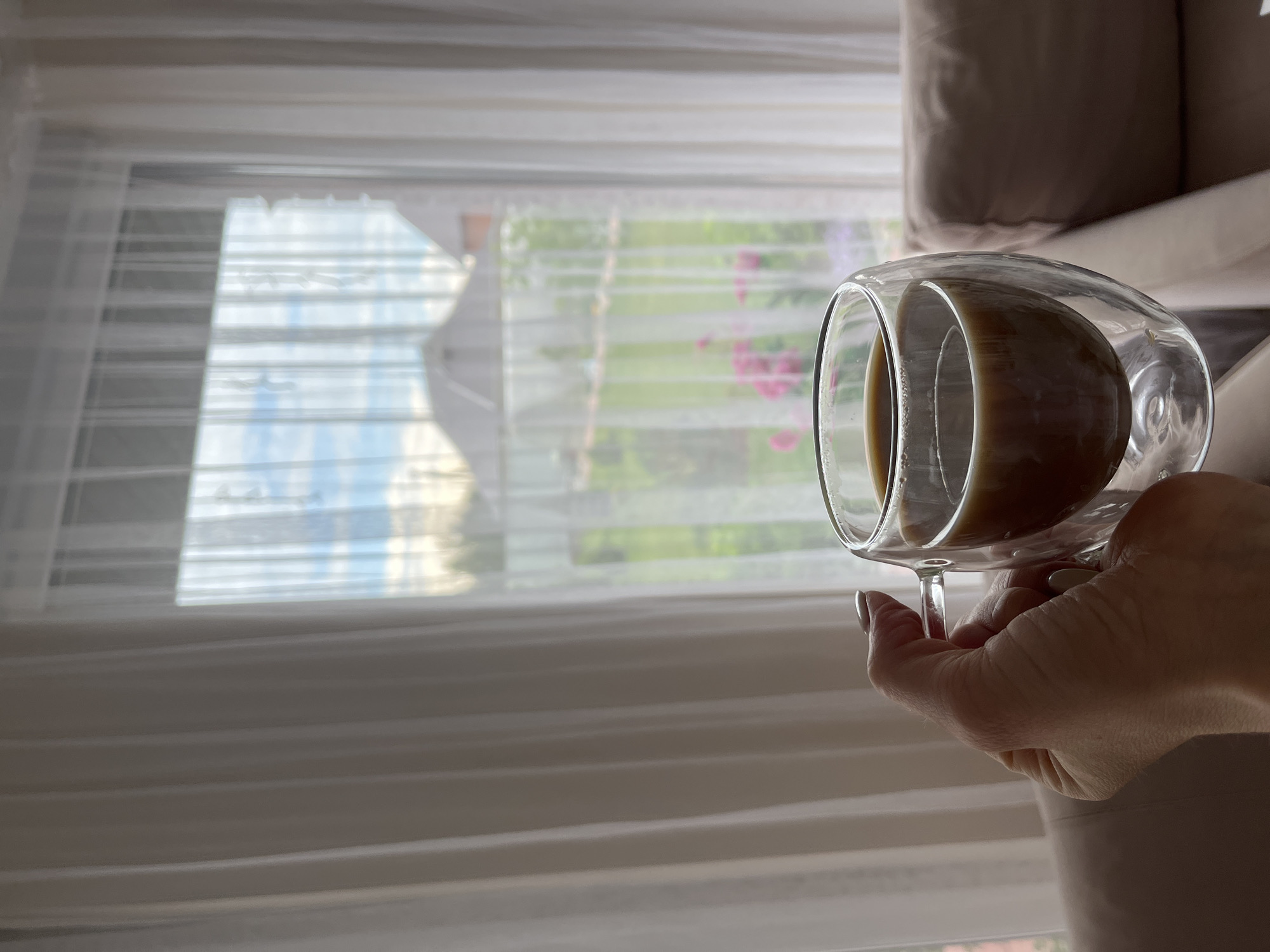 A woman holds a double-walled glass cup filled with a light-colored liquid, likely coffee with milk or a latte. The view through the window behind her suggests a residential area, with a building in the distance and greenery visible, potentially indicating a location in Chaikovsky, Russia, although that cannot be definitively determined from the visual information alone. The lighting is soft, possibly from an overcast day. The interior suggests a domestic setting, with a couch or other upholstered furniture partially visible. The scene appears calm and peaceful.