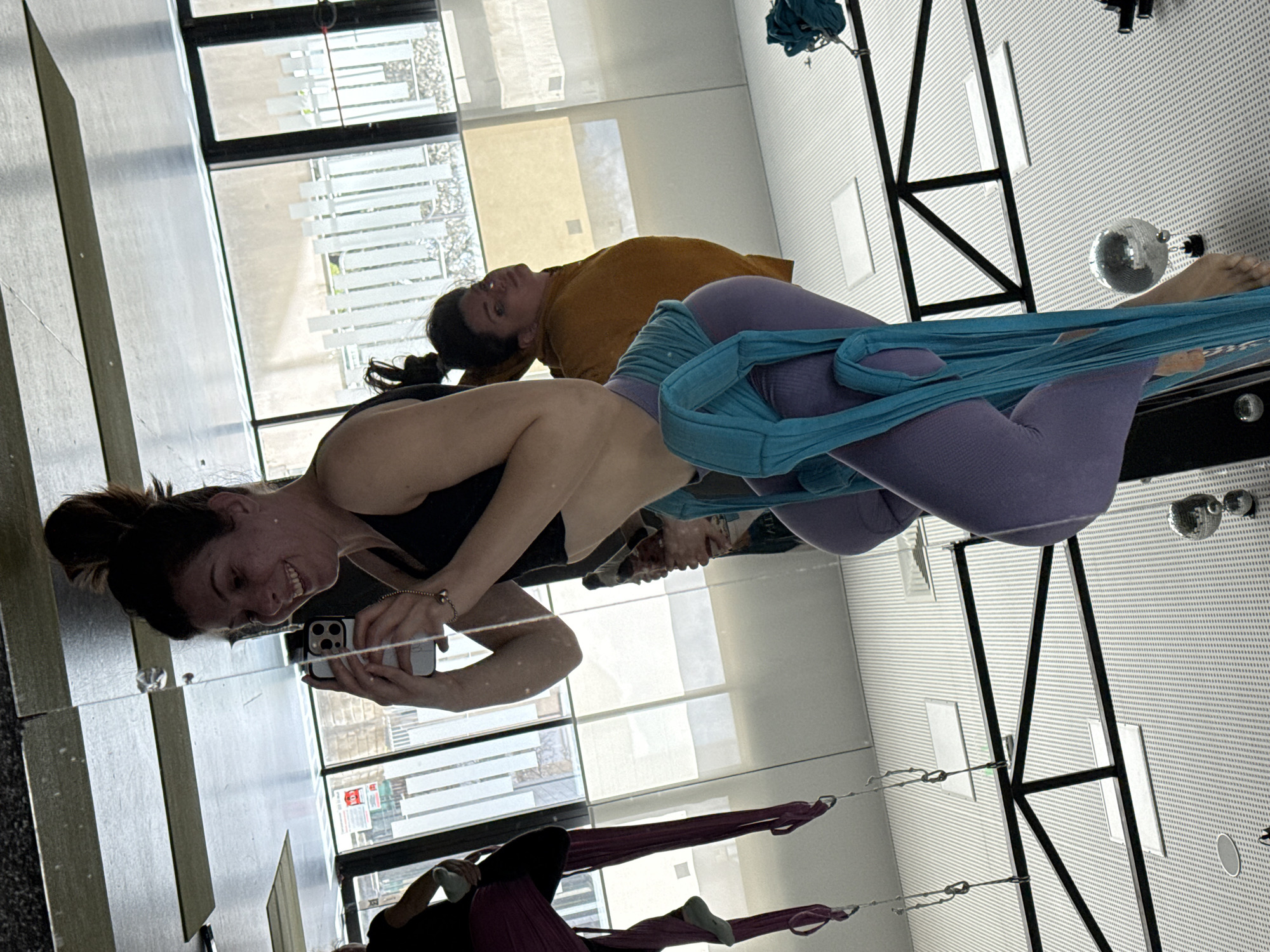 Here's a detailed description of the image:
**Scene:** An indoor space, likely a yoga or aerial fitness studio. The perspective suggests the photo is taken from a mirror reflection.
**People:** Two women are visible in the foreground.
* One is holding a phone and taking a picture in the mirror. She is wearing a black sports bra.
* The other is participating in aerial fitness using a blue hammock. She is wearing lavender leggings.
* There are people in the background with orange and purple hammocks.
**Objects:** Aerial hammocks, a mirrored surface, a cell phone, fitness attire, windows, a disco ball.
**Actions/Interactions:** One woman is taking a selfie in the mirror while the other is engaged in aerial yoga or fitness.
**Environment:** The location appears to be indoors near windows, allowing natural light. The ceiling has a patterned or textured surface. The location in the city of Unknown, Unknown.