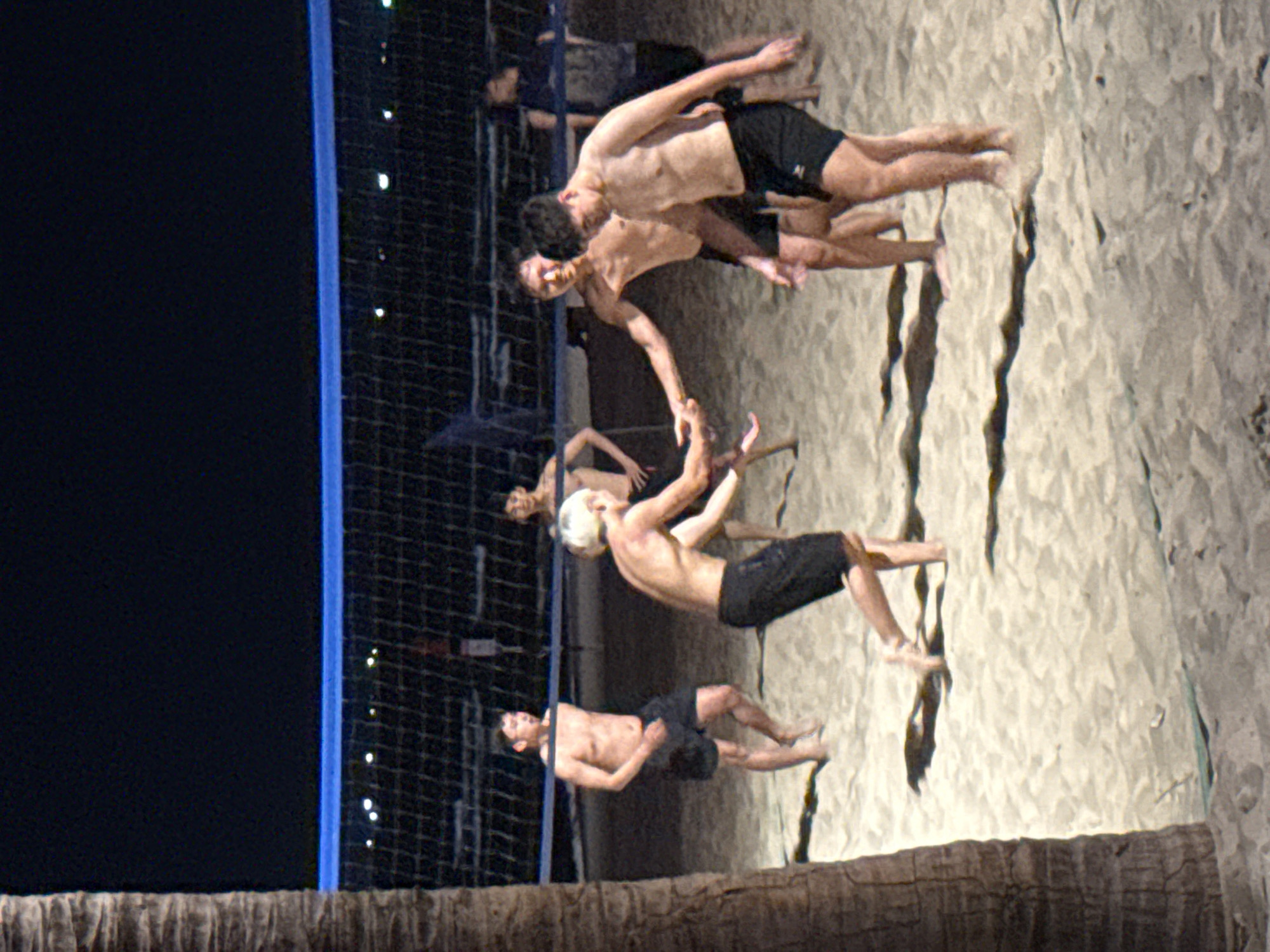 The image depicts a beach volleyball game at night. Six shirtless men wearing shorts are playing on a sandy court, with a net visible on the left side of the frame against a dark sky. The foreground appears to be the top of a palm tree trunk. While Đà Nẵng, Vietnam is known for its beaches, confirmation of this specific location requires further details or contextual clues within the image that identify it as such.
