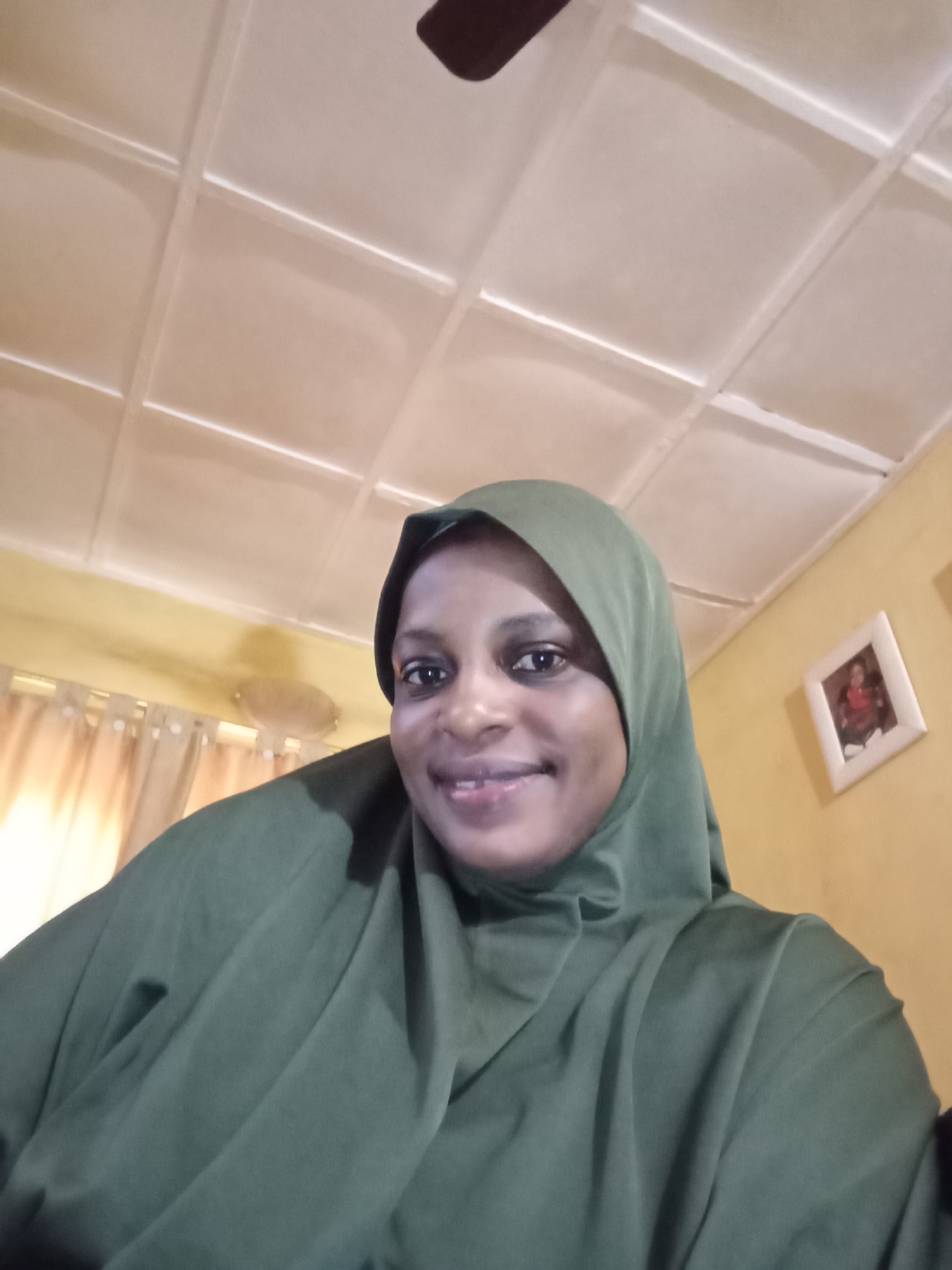 A woman wearing a green hijab smiles at the camera indoors. The room has a cream-colored ceiling with a grid pattern and yellow walls. A framed picture hangs on the right wall. Curtains are visible in the background. The image is a selfie.