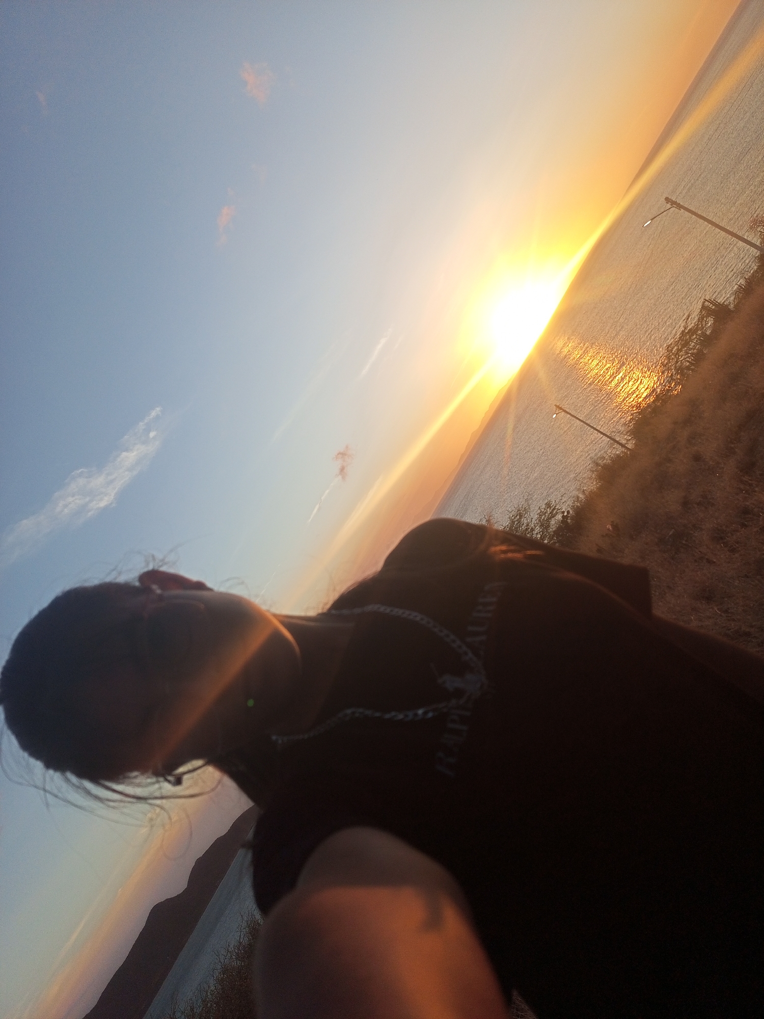 This image captures a sunset scene over a body of water, located in Juan Griego, Venezuela. The composition is severely tilted, with the horizon line running diagonally from the lower left to the upper right.
In the foreground, a person's upper body is visible, largely silhouetted against the bright sunset. Their head, turned slightly to the left, shows dark hair. A silver-colored chain or necklace is worn around their neck, and a dark-colored shirt with faint white text, possibly "RALPH LAUREN," is visible on the chest. A lens flare artifact, appearing as a green dot, is present near the person's right ear. Part of an arm or hand, illuminated by warm light and out of focus, occupies the bottom right corner of the frame, suggesting a selfie or close-up shot.
The sun, a dominant bright source, is positioned along the diagonal horizon in the upper right quadrant, casting intense light across the scene and reflecting off the water's surface in shimmering gold and orange hues. The sky transitions from a light blue with sparse, wispy clouds in the upper left to a brilliant orange and yellow near the sun.
Across the water, a distant, dark landmass, likely mountains or hills, is silhouetted against the sky. On the immediate right side of the frame, a sloping terrain covered in dry, sparse vegetation descends towards the water. Two dark, slender poles, resembling streetlights or utility poles, extend from this land into the water's edge. The top of one pole shows a visible light source. The water appears calm with rippled textures due to the sun's reflection.