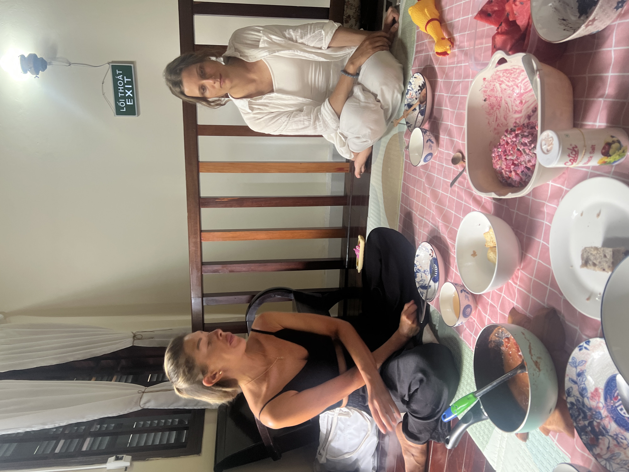 Two women are sitting on the floor, which is covered with a pink and white checkered mat, in a room in Đà Nẵng, Vietnam. The woman on the left is wearing a black tank top and black pants. The woman on the right is wearing a white top and white pants. They are both looking at the camera. There are several bowls of food on the mat, as well as a can of salt and a yellow rubber chicken toy. A wooden railing is visible behind the women, and a sign that reads "LỐI THOÁT EXIT" is on the wall. A window with white curtains is to the left of the frame. The lighting appears to be artificial, possibly from a ceiling lamp.
