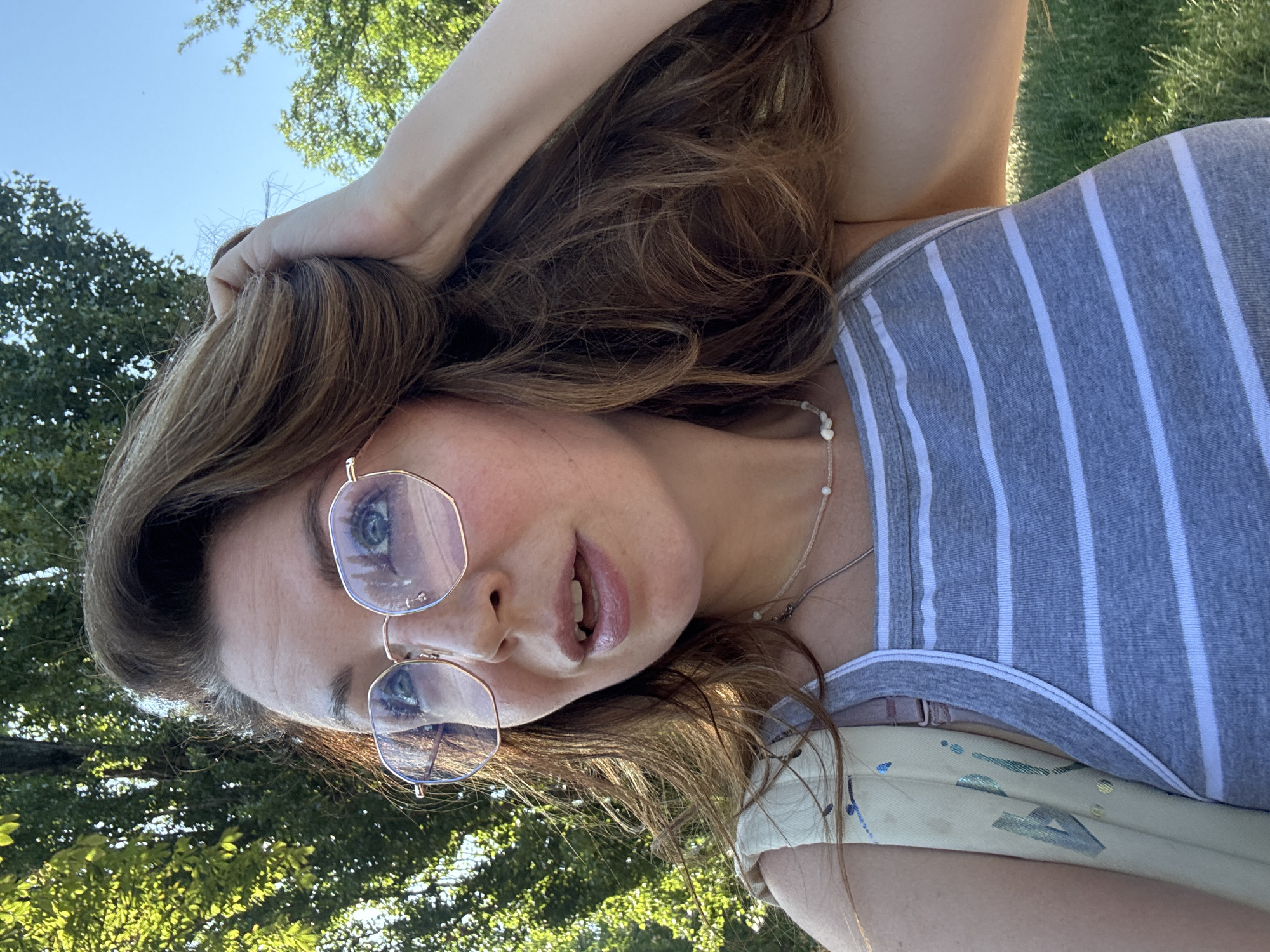 The media file is a close-up, portrait-style photo of a young woman outdoors. She has long, wavy brown hair, and is wearing glasses with rose-gold frames. Her eyes are blue-green and she has on a grey and white striped tank top. Around her neck, she wears a delicate necklace with small white beads. The background consists of green foliage and a bright blue sky, suggesting it's a clear day. The lighting indicates it's likely daytime, possibly late morning or afternoon. The woman's hand is visible, resting on her head and in her hair. Her mouth is slightly open, giving the impression she might be speaking or about to speak. The overall mood of the image appears casual and relaxed. There is no visible text in the image. The location context is Kryvyi Rih, Ukraine.