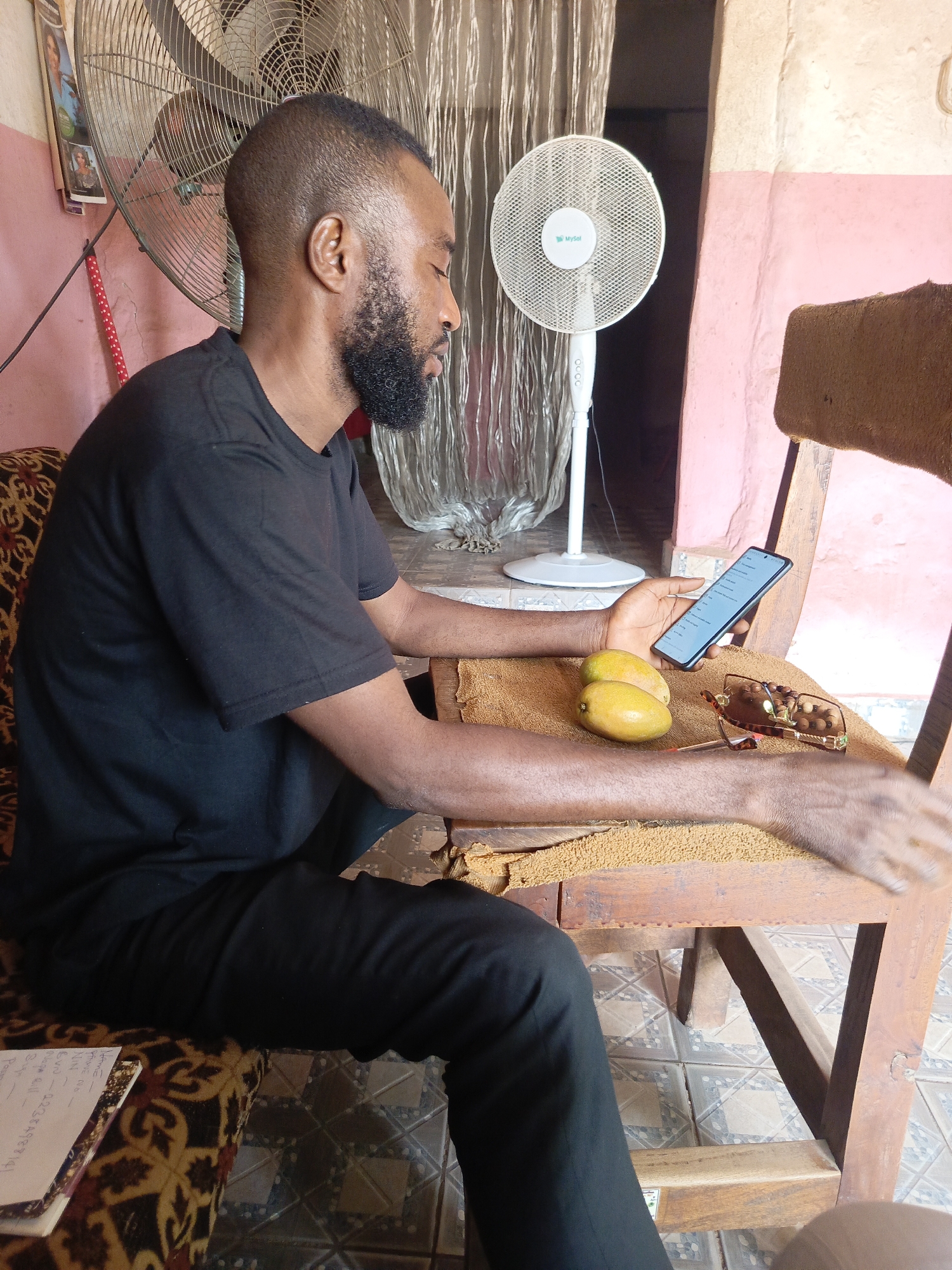 A man with a beard is sitting on a wooden chair in a room. He is wearing a black t-shirt and black pants. He is holding a smartphone in his right hand and looking at the screen. There are two mangoes and a pair of sunglasses on the chair next to him. There are two fans in the background. The location appears to be inside a house, possibly in the city of Unknown, Unknown.