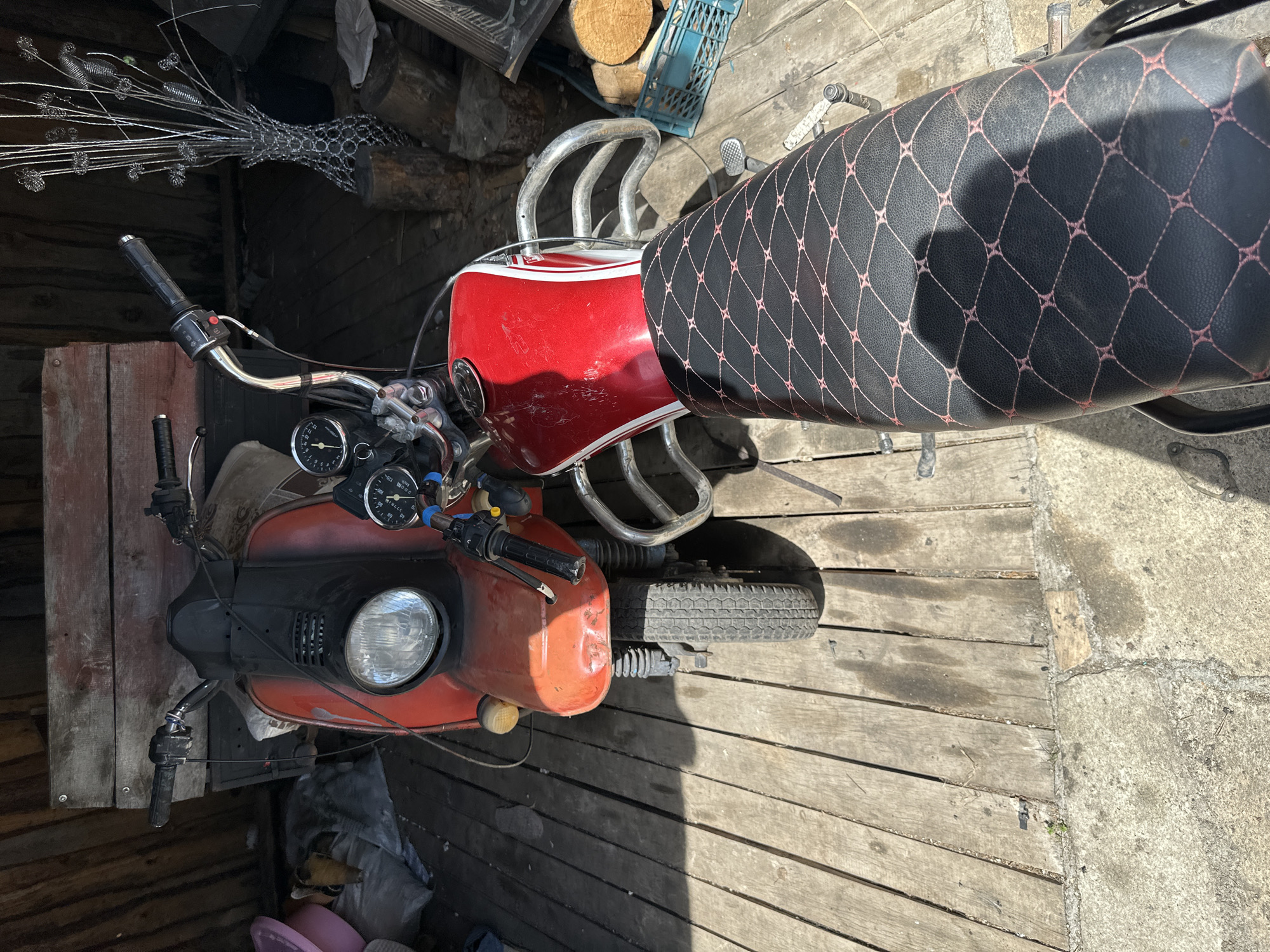 The image shows two vintage motorcycles parked on a wooden deck. The motorcycle in the foreground has a black seat with a diamond-pattern stitching in pink. The motorcycle in the background is orange and black. Both motorcycles appear to be in a state of disrepair. The environment appears to be an outdoor shed or garage, with wooden walls and a concrete floor. There are various objects in the background, including logs, a blue plastic crate, and a metal sculpture of flowers. The lighting suggests it is daytime, with sunlight casting shadows on the ground. The scene is static, with no people or discernible action. The location is unknown, as is the city.