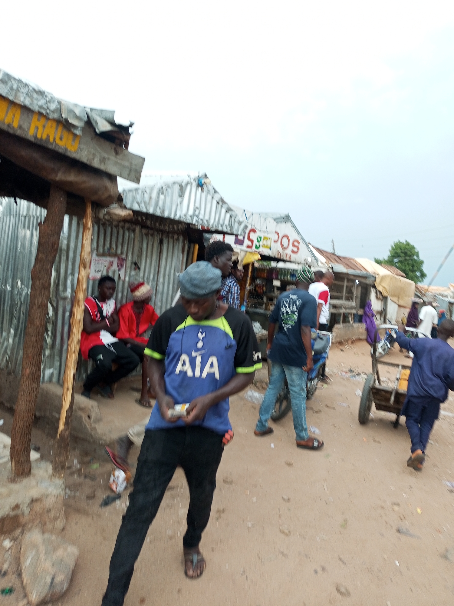 The image captures a street scene in Gombe, Nigeria, with a focus on several individuals and makeshift structures. In the foreground, a person wearing a blue t-shirt with "AIA" and a Tottenham Hotspur logo, along with black pants and sandals, is visible. They are holding what appears to be a cigarette pack. To their left, two men are seated, one in a red shirt and the other in a red and white patterned attire. Behind them, a corrugated metal structure serves as a shop or shelter, with a wooden beam and a sign that reads "MA HAGGU" in yellow lettering.
Further down the street, other individuals are present. A person in a blue t-shirt and jeans is walking away from the camera, near a motorcycle. Another individual in a blue jacket is pushing a cart with a wheel. Several other structures, seemingly shops with corrugated metal roofs and tarpaulin coverings, line the street. The ground is dirt, with scattered debris. The overall atmosphere suggests a bustling, informal marketplace or residential area. The sky is overcast.