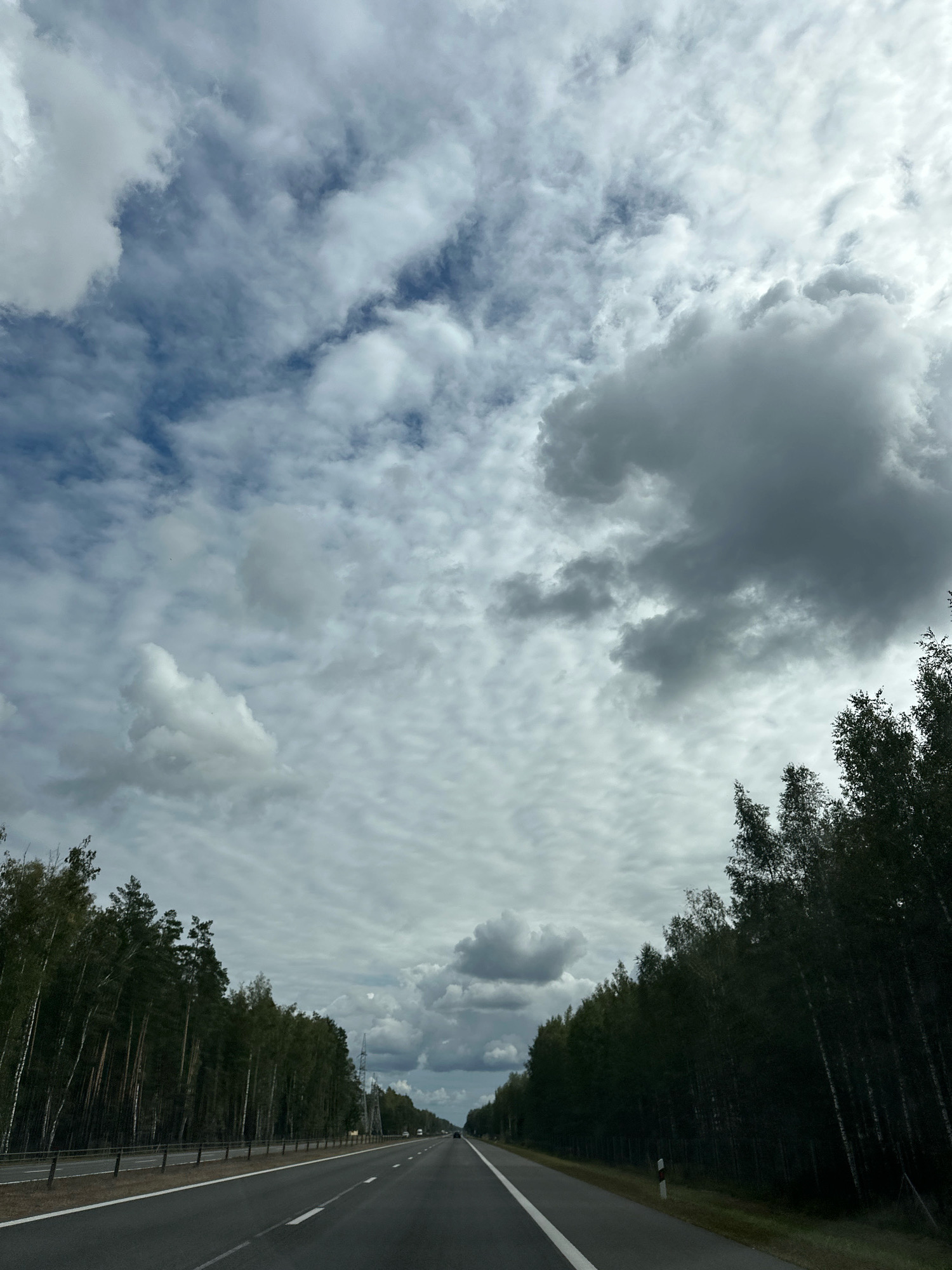 The image depicts a view from a vehicle traveling on a multi-lane highway. The highway is flanked by dense deciduous and coniferous forests on both sides. The sky is filled with a dynamic arrangement of clouds, ranging from light, scattered formations to heavier, darker masses, with patches of blue sky visible. The lighting suggests it is daytime, possibly overcast or with intermittent sun. No people or other vehicles are clearly visible in the immediate foreground or along the sides of the road, though distant vehicles can be seen on the highway ahead. The overall scene suggests a journey through a rural or semi-rural area in Belarus, consistent with locations like Лапіцкі сельскі Савет.