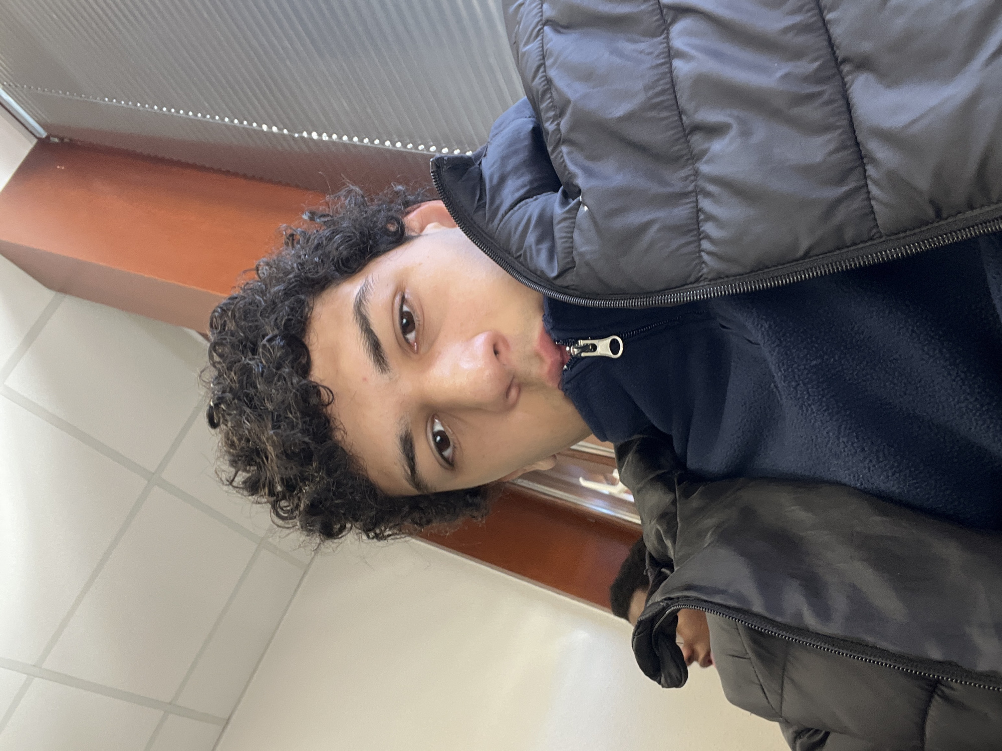 A young man with curly black hair and brown eyes is facing the camera. He is wearing a dark blue fleece jacket and a black puffer jacket over it. He is pouting his lips slightly. The background consists of white square tiles and a light brown horizontal surface, possibly a shelf or counter. The location is unknown.
