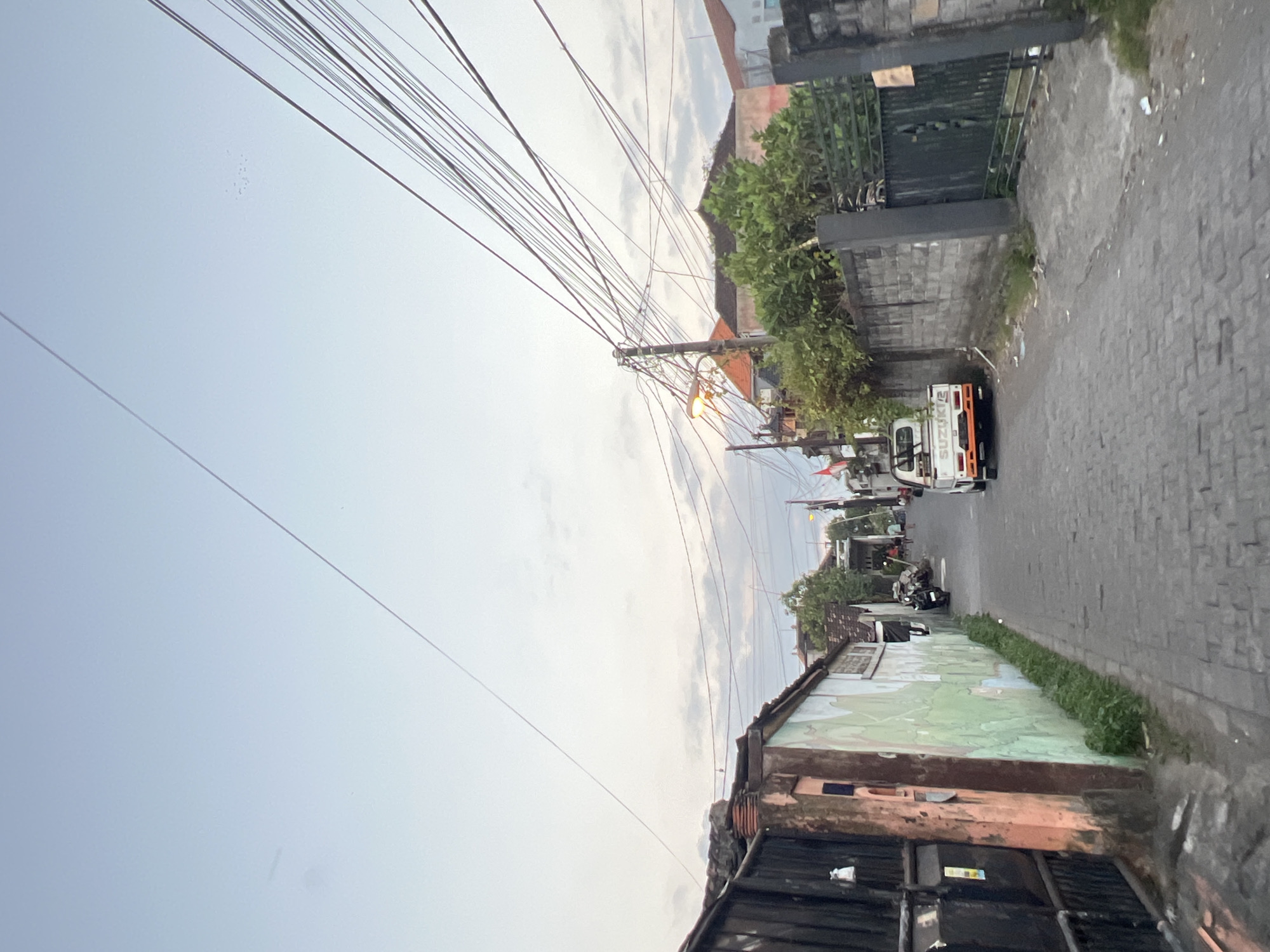 The image shows a narrow, paved street in Denpasar, Indonesia, with buildings and walls on either side. A white Suzuki pickup truck is parked on the right side of the street. Overhead, a dense network of electrical wires crisscrosses the sky. The street is lined with vegetation and appears to be in a residential or mixed-use area. The sky is overcast with scattered clouds, suggesting it might be late afternoon or early evening. The overall impression is of a typical urban street scene in Southeast Asia.