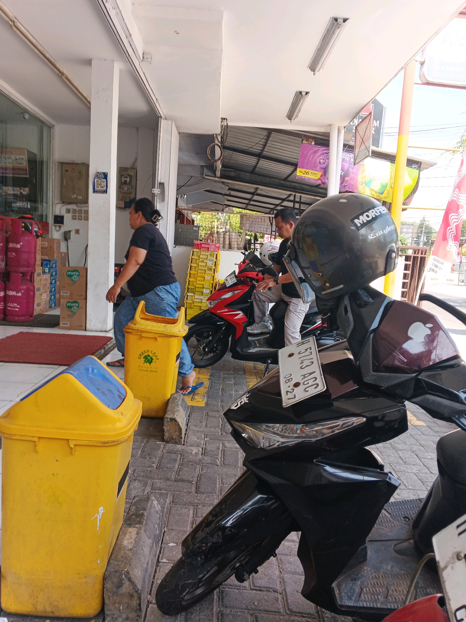 The image depicts an outdoor scene in front of a store in Kalirejo, Indonesia. In the foreground, a black motorcycle is parked, with its front wheel and part of its fairing visible. A license plate, partially obscured, reads "H 5713 AGC 08.27". To the left, a yellow trash can with a blue lid stands next to a gray curb. Further back, another yellow trash can is visible, with a woman in a black t-shirt and blue jeans sitting on its edge, wearing blue flip-flops. She is looking towards the right. Beside her, a red motorcycle is parked, and a man in a black t-shirt and light-colored pants is seated on it, facing left. He is wearing a black helmet with "MORFE" and "Calygloss" stickers on it. Behind the man, another helmet is visible. The store entrance is to the left, with shelves of bottled water and pink propane tanks visible through the glass. Yellow crates are stacked to the right of the store. Various signs and posters are displayed above the storefront. The ground is paved with interlocking gray bricks, with yellow markings indicating parking spaces. The overall lighting suggests it is daytime, with sunlight casting shadows.