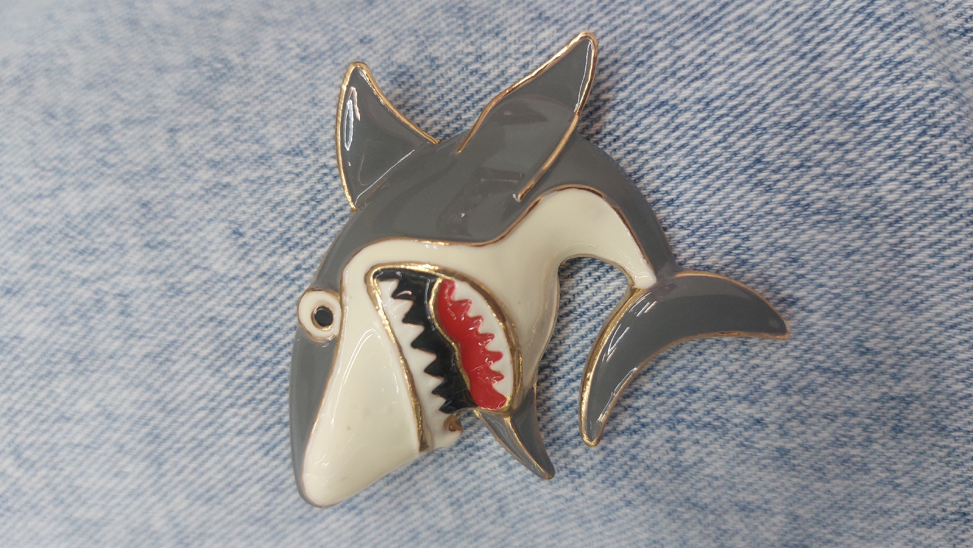 A close-up image shows a shark-shaped brooch pinned to a light blue denim surface. The brooch is made of enamel in gray, white, red, and black with a gold-colored outline. The shark is stylized and shows an open mouth with red gums and black teeth. The background is the textured weave of the denim fabric.