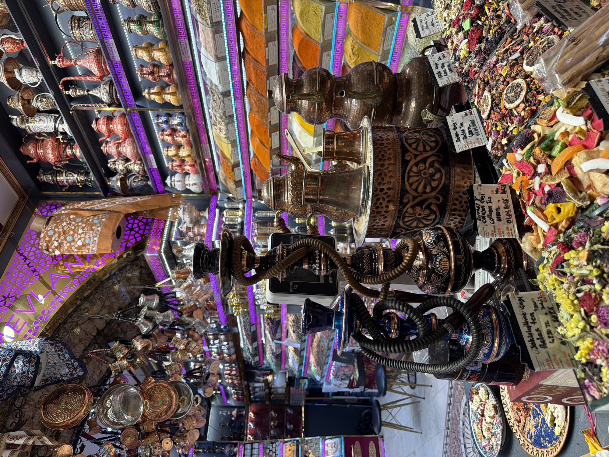 The image captures a vibrant display of goods within a marketplace in Istanbul, Turkey. The scene is dominated by traditional Turkish items, including numerous copper teapots and samovars displayed on shelves, many with intricate designs and in various sizes. In the foreground, a large, ornate hookah pipe stands prominently, its dark metal base and winding hose suggesting its decorative or functional purpose.
To the right of the hookah, several stacked copper vessels, likely for brewing coffee or tea, are visible. Behind these, shelves are laden with an array of spices and dried fruits, meticulously arranged in neat rows. The colors range from deep reds and oranges of paprika and chili powder to the earthy tones of other spices, interspersed with the vibrant hues of dried fruits like apricots, figs, and citrus slices. Small price tags are attached to some of the products, indicating that this is a retail environment.
The lighting in the market is a mix of warm, ambient light and striking purple neon accents that line the shelves and archways, creating a lively and somewhat theatrical atmosphere. The ceiling features a traditional arched design, adorned with decorative elements. Hanging from the ceiling in the upper left of the image are various copper bowls and utensils, adding to the rich texture and visual density of the market.
The overall impression is that of a bustling Turkish bazaar, possibly the Grand Bazaar or the Spice Market (Egyptian Bazaar), renowned for their eclectic mix of traditional crafts, spices, and food items. The presence of the hookahs and the abundance of spices and dried fruits strongly points to this type of marketplace. No people are clearly visible in the frame, but the setup implies active commerce. The floor is covered with a patterned rug, further contributing to the traditional aesthetic.