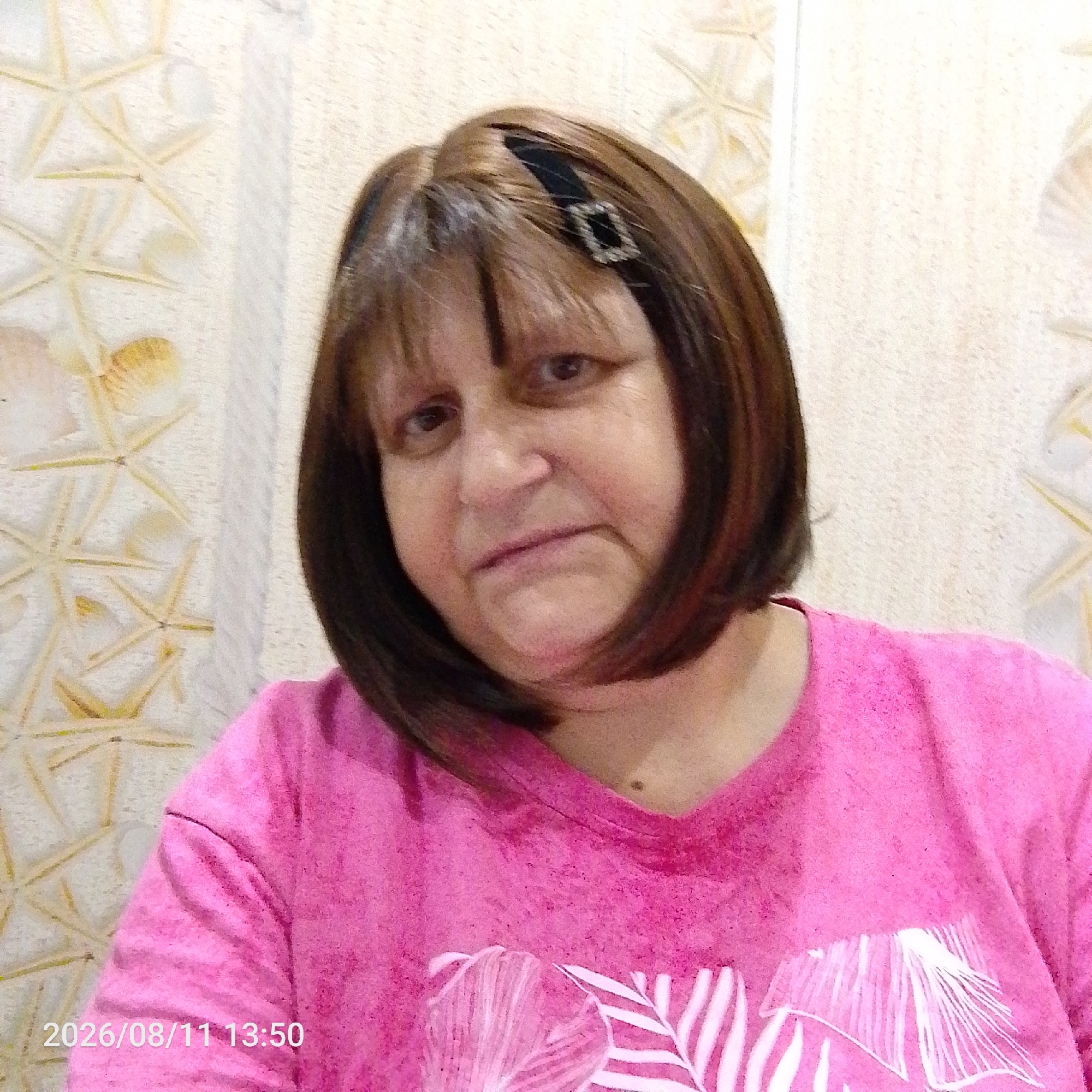 A medium shot photo shows a woman from the chest up, facing the camera and slightly to her right. She has brown hair with lighter highlights styled in a bob, reaching just below her chin, and bangs. She is wearing a bright pink t-shirt with a subtle white palm leaf design. A black hair accessory with a small, rectangular, jeweled ornament is visible in her hair. The background is a wall with a light-colored, textured wallpaper featuring a repeating pattern of starfish and shells. A timestamp in white text, "2026/08/11 13:50", is visible in the bottom left corner of the image.