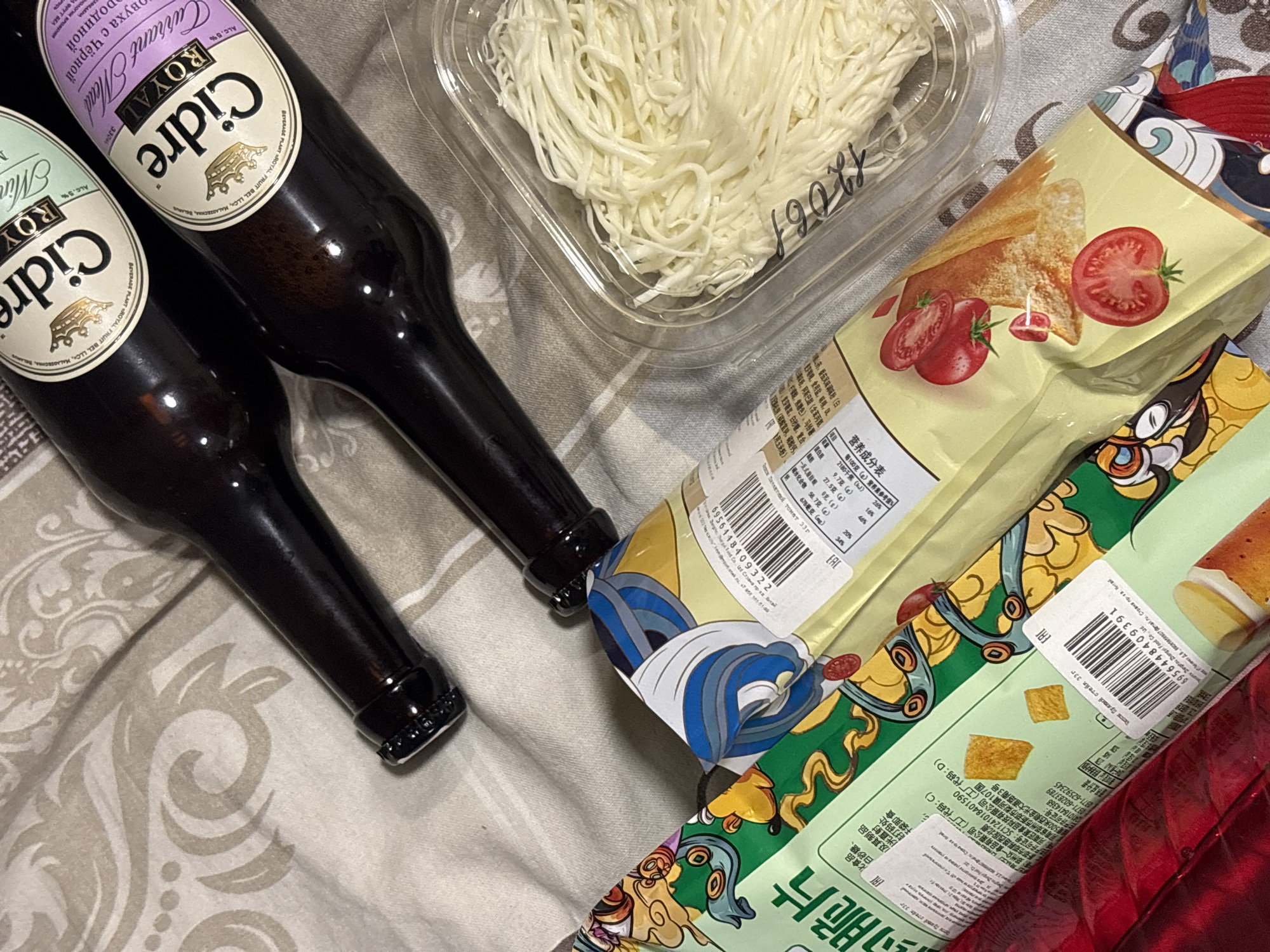 The image shows a still life on a patterned surface. There are two dark glass bottles of "Cidre Royal," one "Currant Mead" and one "Mint" variety. Next to the bottles is a clear plastic container filled with string cheese. Adjacent to the cheese are two bags of chips, one tomato flavored and one "spicy steak." The text on the chip bags is partially in Chinese. The location is not clear from this image.