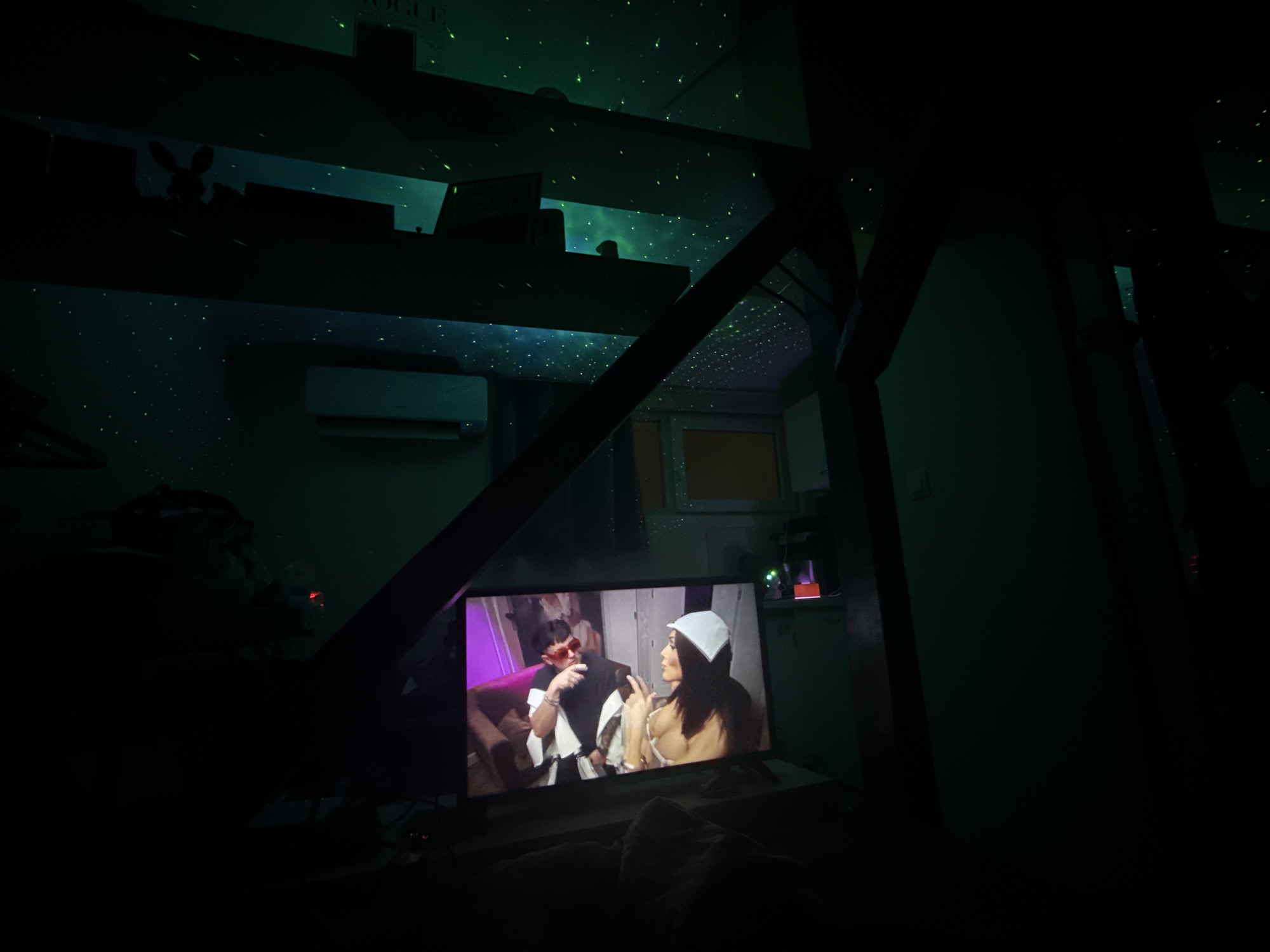 The image shows a dimly lit room in Sofia, Bulgaria. The focal point is a television screen displaying two individuals, one with red sunglasses and the other wearing a white head covering. The room has shelves with various items and an air conditioning unit on the wall. The ceiling is illuminated with a pattern of small, bright dots, resembling stars. Furniture or other objects are partially visible in the foreground.