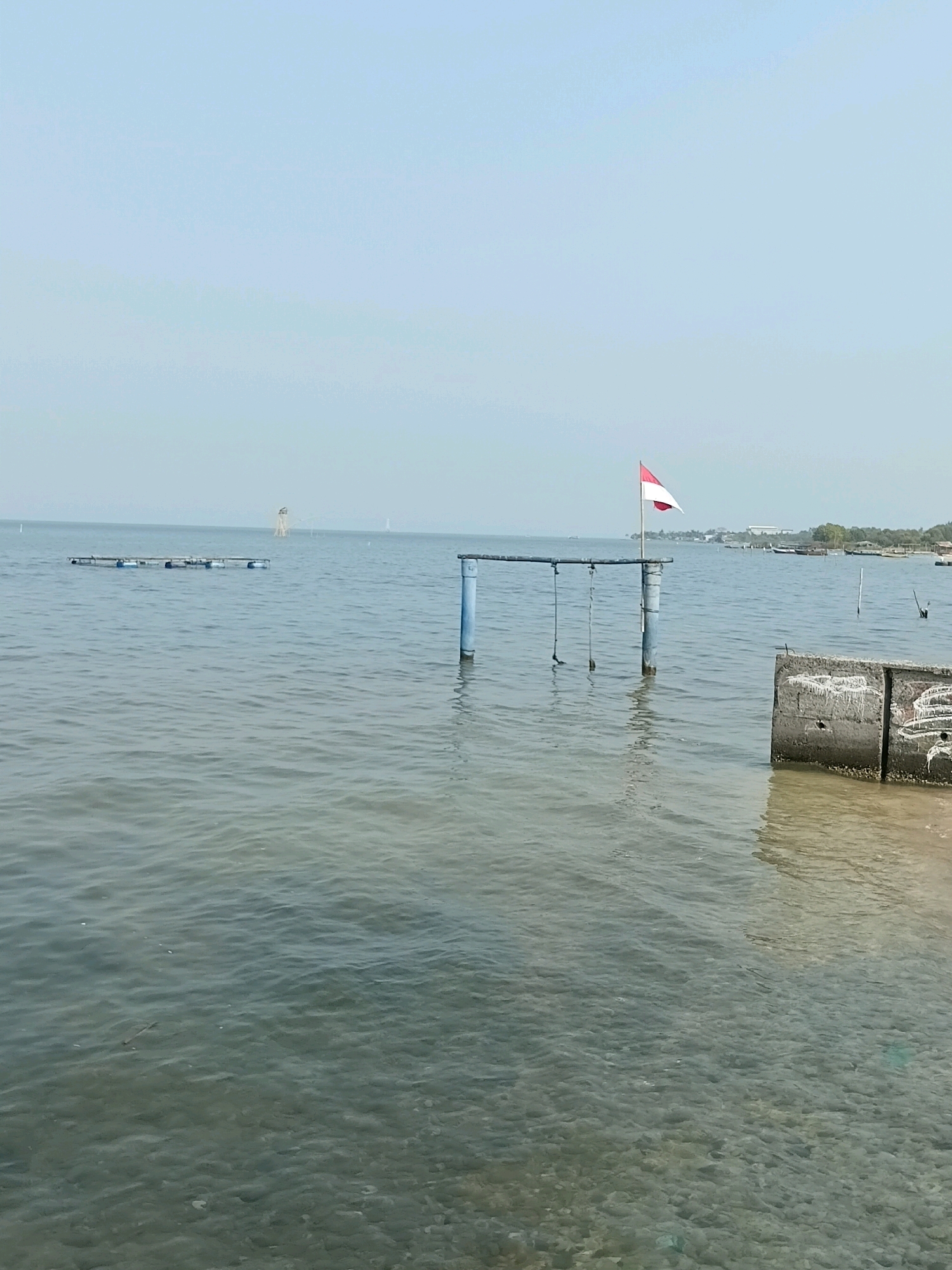 The image depicts a calm body of water, likely a bay or inlet, in Cikal, Indonesia. The water is clear enough to see the rocky bottom in the foreground. Several man-made structures are visible in and around the water. In the center of the image, a blue wooden pole stands in the water, supporting a horizontal beam from which hang three ropes with objects at the ends. A red and white Indonesian flag is flying from another blue pole on the right side of this structure. To the left, a rectangular floating platform with blue barrels attached is anchored in the water. Further in the distance, a taller, narrow structure, possibly a lighthouse or observation tower, is visible, along with other boats and a hazy coastline with trees and buildings. The sky is a pale blue and overcast, suggesting a clear but possibly humid day. The overall scene is tranquil, with a focus on the intersection of the natural environment and human infrastructure.