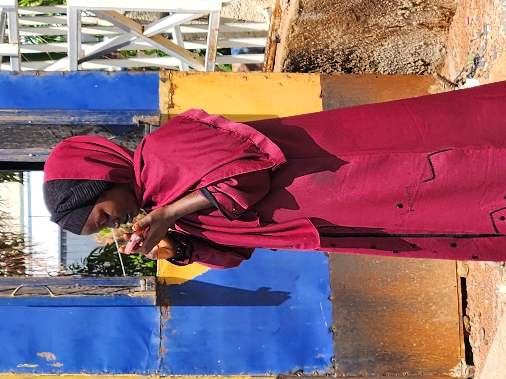 A young Black woman wearing a burgundy hijab and a matching long dress stands outdoors in the city of Jos, Nigeria, looking down at a pink object she is holding in her hands. The woman is positioned in front of a weathered metal structure painted in sections of bright blue and yellow. A white wooden gate or fence is visible to her right, and beyond that, some greenery and a rocky embankment. The ground is covered with loose gravel and small stones. The woman's head is covered by a dark-colored hijab, and her face is partially obscured as she focuses on the object in her hands. Her dress is loose-fitting and appears to be made of a flowy fabric, with black detailing on the cuffs and down the front. The overall lighting suggests it is daytime.