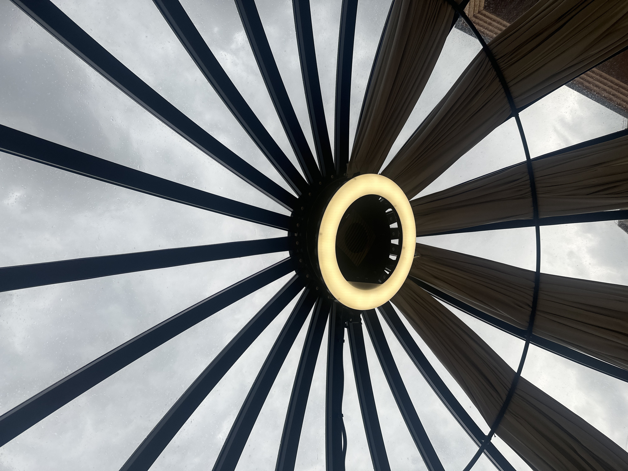 The image shows an upward view of a gazebo-like structure. At the center is a circular light fixture. The structure has a radial pattern of beams extending outwards from the center, with what appear to be cloth panels between some of the beams. The background is an overcast sky, possibly rainy. The style of the gazebo, particularly the combination of beams and cloth panels, may be associated with outdoor event spaces or upscale patios. While the location in Votkinsk, Russia is given, there are no contextual clues in the image itself that directly confirm this location.