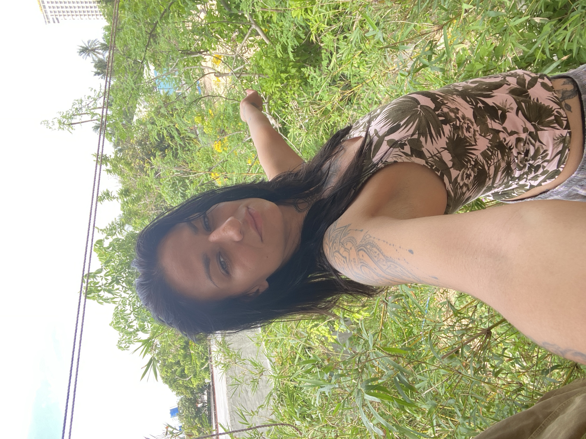 A woman with dark hair and tattoos on her arm and shoulder is taking a selfie outdoors in a green, overgrown area. She is wearing a pink and green floral tank top and grey shorts. She is pointing her right arm out towards the right side of the frame. In the background, there are trees, bushes, and some yellow flowers. A tall white building with many windows is visible in the distance, as are palm trees. The sky is overcast. This scene is located in Mui Ne, Vietnam.