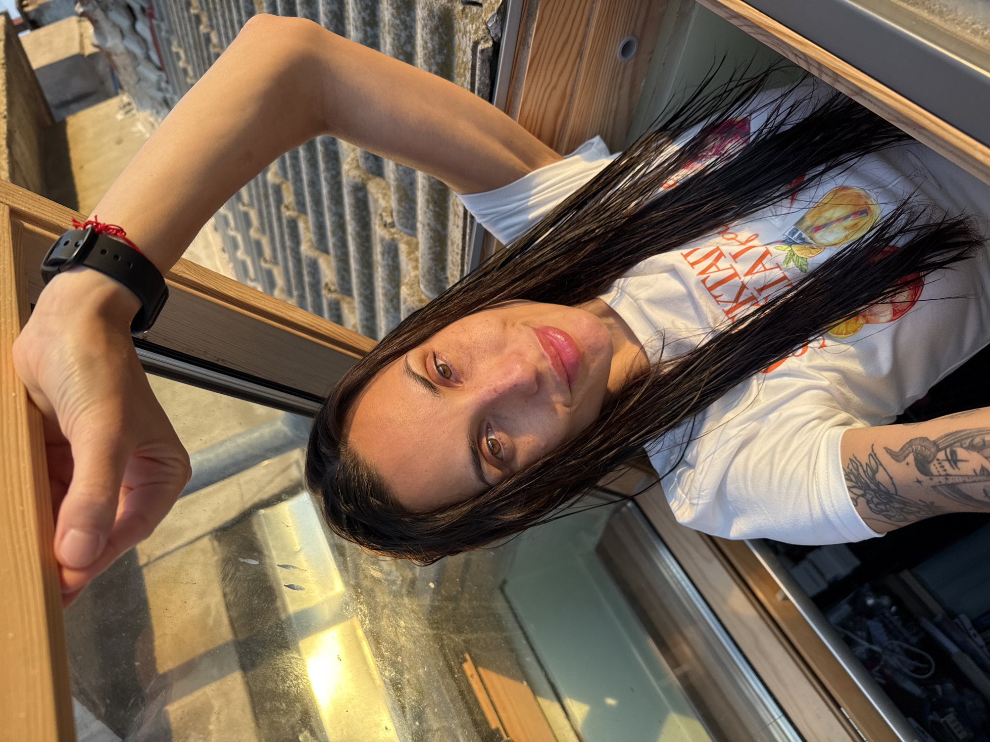 A young woman is captured in a selfie-style portrait, looking directly at the camera with a slight smile. She appears to be emerging from or leaning into a skylight or window opening, with her body oriented horizontally due to the camera's angle. Her left arm is extended and bent, resting on the wooden frame of the opening, and she is wearing a black watch with a red string bracelet on her wrist. Her right arm is bent, revealing a detailed black and white tattoo on her forearm, possibly depicting a horned creature and floral elements.
She has long, dark, straight hair that falls across her shoulders and chest. She is wearing a white short-sleeved t-shirt with a graphic design featuring colorful fruit illustrations and text. The visible text on her shirt reads "VINTAGE FRUIT ATLANTA".
The lighting suggests a golden hour, with warm sunlight illuminating her face and the dirty glass pane of the window. In the background, beyond the window, a corrugated metal roof is visible, suggesting an exterior urban or semi-urban setting. The overall scene indicates the photo was taken from an elevated position, possibly from an attic or upper floor looking out over rooftops in Pleven, Bulgaria.