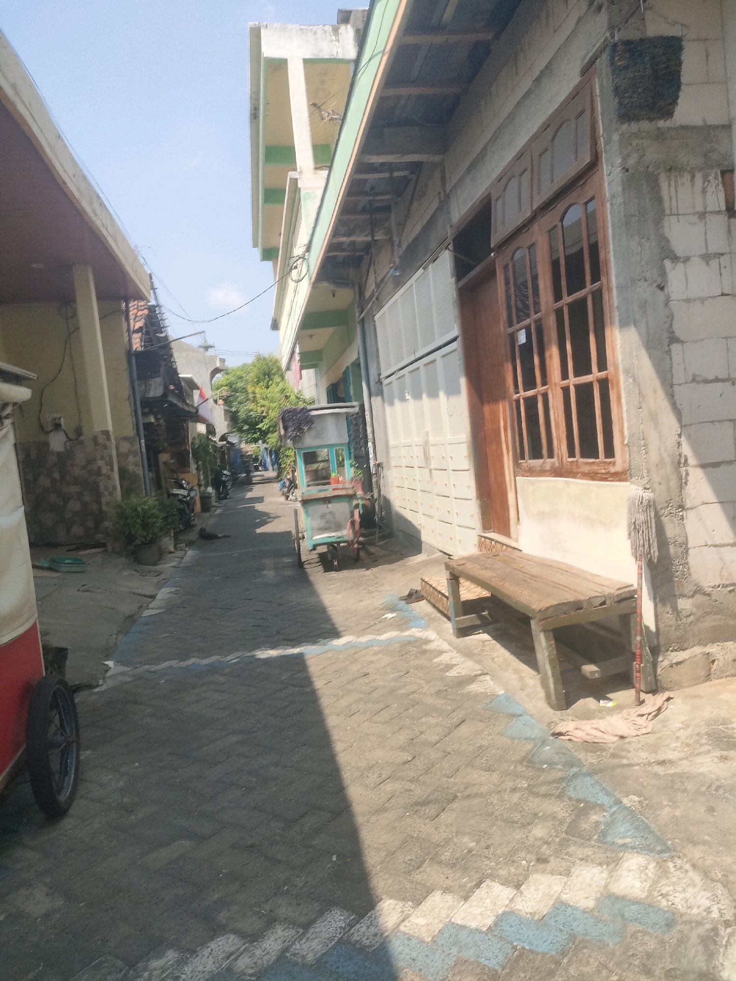 The image depicts a narrow alleyway in a residential area, likely in Surabaya, Indonesia, characterized by buildings of varying construction and age. The foreground features a weathered wooden bench placed on a cobblestone path marked with alternating blue and white rectangular patterns. To the left, a portion of a red and white vehicle, possibly a food cart, is visible with its wheel. A mop leans against the wall beside the bench.
The middle ground of the alley is occupied by a small, mobile food cart with a teal-colored body and a glass display case. Behind the cart, parked motorcycles suggest local transportation. The alley is lined with buildings constructed from concrete blocks and plastered walls, some with exposed brickwork. One building has a wooden door and a large, multi-paned window. Further down the alley, greenery from trees and potted plants adds a touch of nature to the urban landscape. The sky above is clear and blue, indicating a sunny day. The overall impression is of a typical, bustling urban environment in Indonesia.