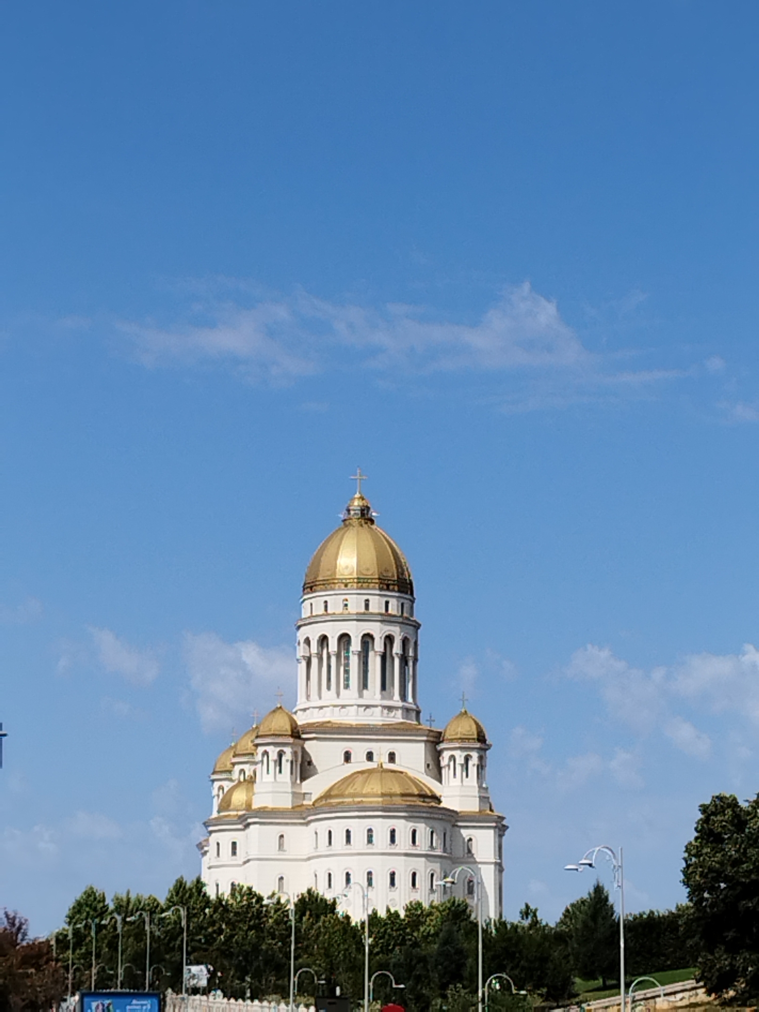 The image shows a large, white cathedral with multiple golden domes against a clear blue sky with scattered white clouds. The cathedral has a tall central dome topped with a cross, and several smaller domes surrounding it. The building features arched windows and intricate architectural details. In the foreground, there are lush green trees and streetlights. The scene appears to be outdoors in a city. This is the Cathedral of the Salvation of the Romanian People, located in Bucharest, Romania.