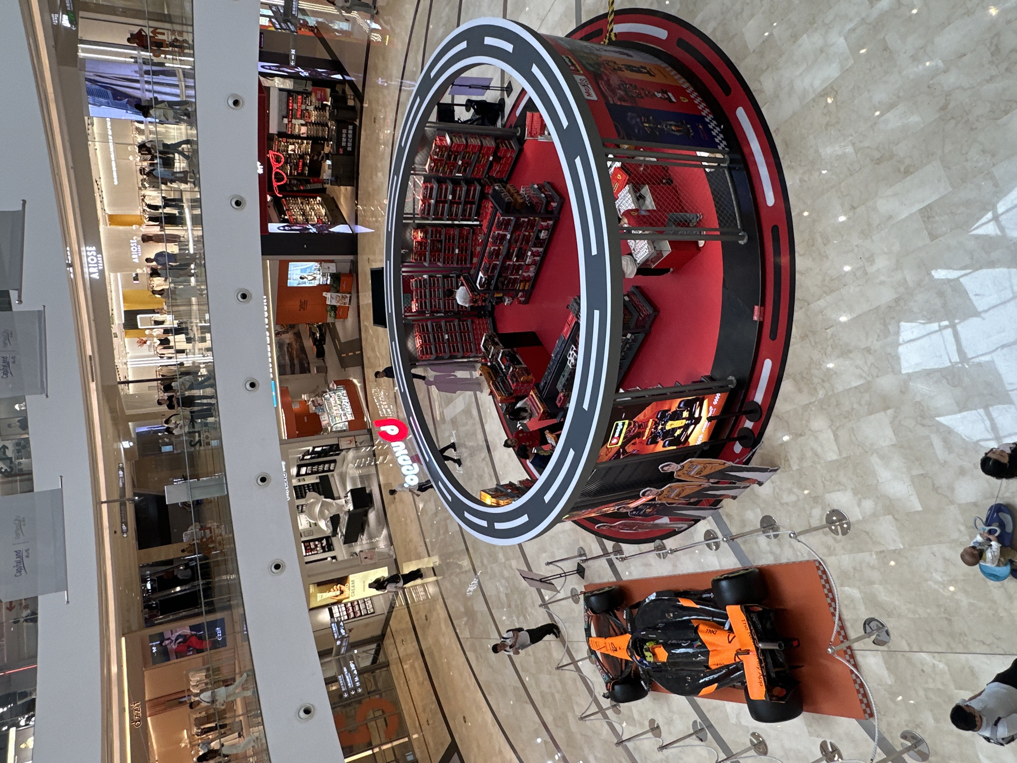 Here's a description of the image:
**Location:** A multi-story shopping mall in Yuzhong District, Chongqing, China.
**Scene:** The image is taken from an elevated perspective, looking down into the mall. The mall features multiple shops along the vertical axis of the photo.
**Objects:**
* A large Formula 1 display. This features a circular structure with shelving units containing items (possibly merchandise). The display includes imagery related to Formula 1 racing.
* A full-scale replica of a McLaren Formula 1 race car, prominently displayed on an orange platform. It is surrounded by stanchions and ropes to prevent access.
* Mall storefronts including Arione, Cream and Dzzit.
**People:**
* People are visible shopping in the mall.
* A few individuals are looking at the F1 car.
**Overall Impression:** The image depicts a typical commercial environment in a shopping mall, with the addition of a display showcasing Formula 1 racing, presumably for promotional or commercial purposes. The location appears modern and well-lit.
