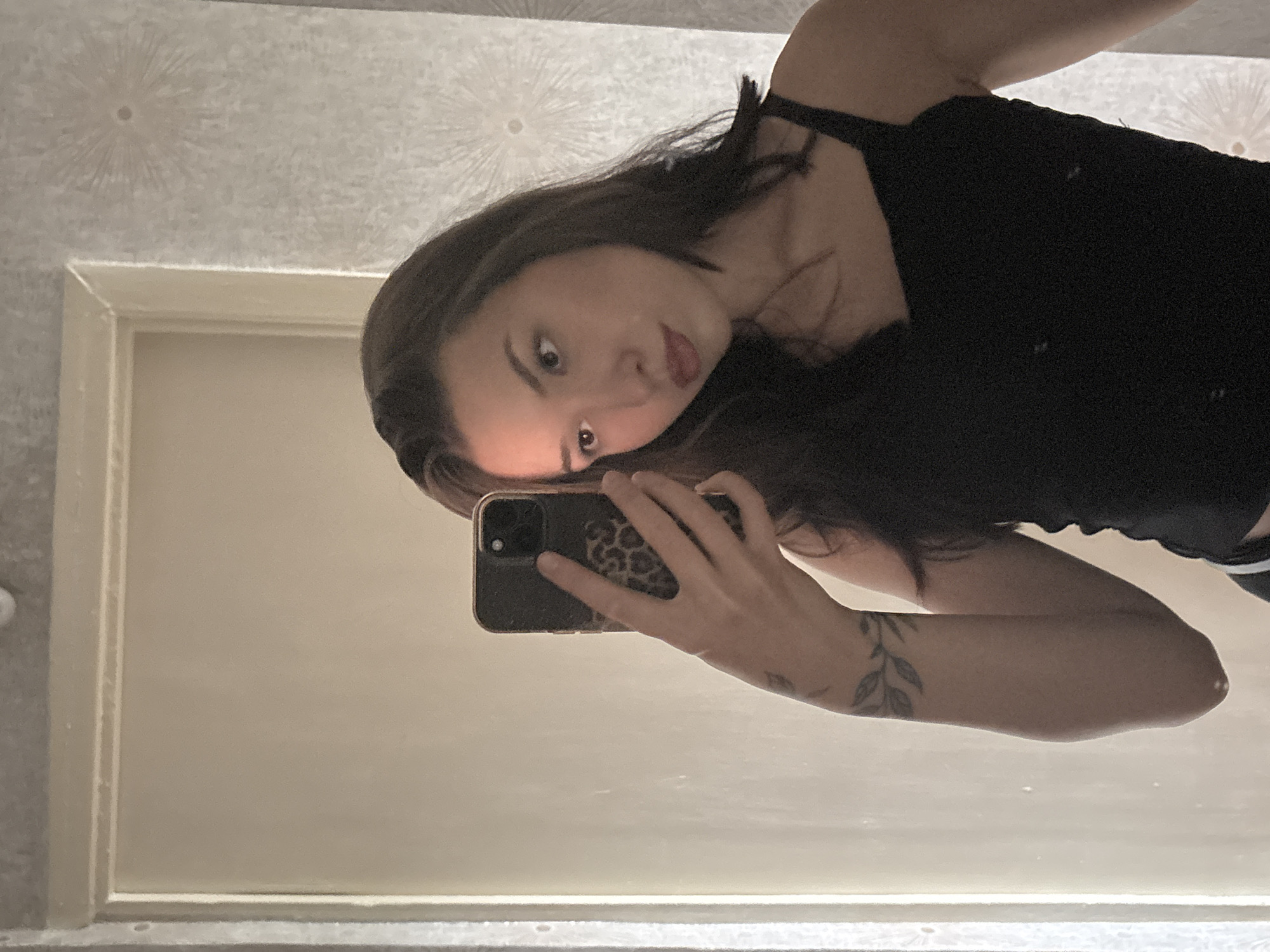 A young woman with dark hair is taking a selfie in a mirror. She is wearing a black tank top. She has a tattoo of a branch with leaves on her left forearm. She is holding a smartphone with a leopard-print case in her right hand. The background consists of a plain wall and a patterned wallpaper. The image does not contain enough information to determine the location in Inskoy, Russia.
