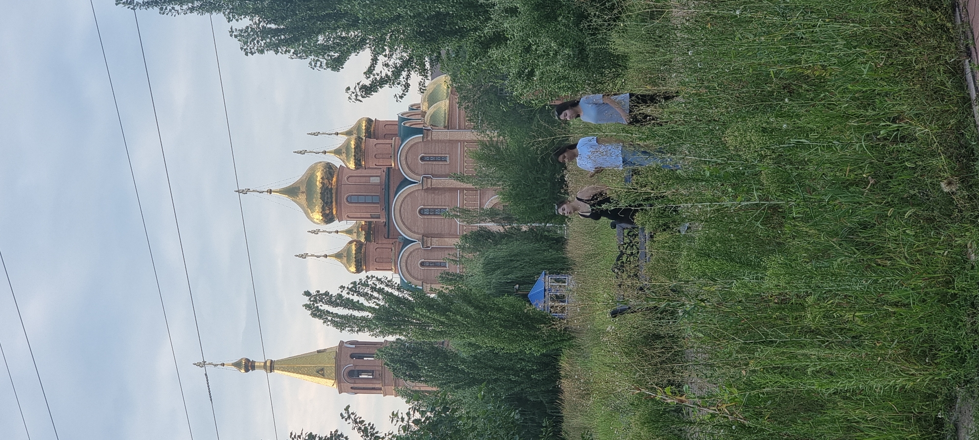The image shows a brick church with golden domes, likely in Aktobe, Kazakhstan. Three young women are standing in a field of tall grass in the foreground, facing the church. The woman on the left is wearing a black tank top and jeans. The woman in the middle is wearing a light blue patterned t-shirt and jeans. The woman on the right is wearing a light blue t-shirt and dark pants. Behind them, a blue gazebo is visible. The sky is overcast. Power lines are visible across the top of the image.