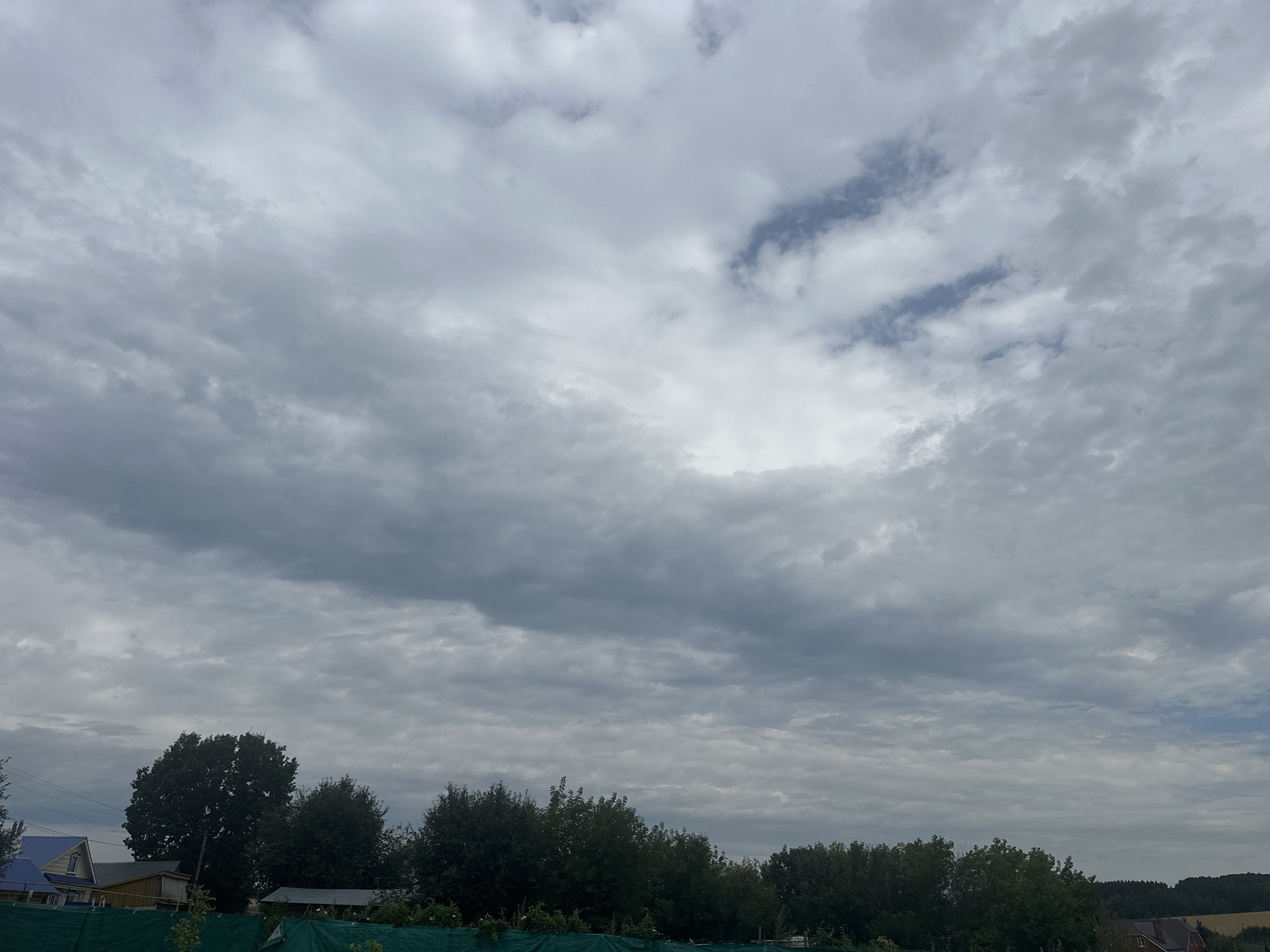 The image captures an overcast sky over a rural or semi-rural area, likely on the outskirts of Улля, Russia. The sky is filled with a dense layer of grey clouds, with some patches of lighter grey and a hint of blue peeking through in the upper right quadrant. Below the sky, a line of dark green trees forms a dense canopy across the middle ground. To the left, several houses with distinct blue and brown roofs are visible, suggesting a residential area. In the foreground, a green fence runs horizontally, partially obscuring what appears to be vegetation or another type of barrier. The overall scene is characterized by muted colors and a sense of diffused light, typical of a cloudy day. No people are visible in the frame.
