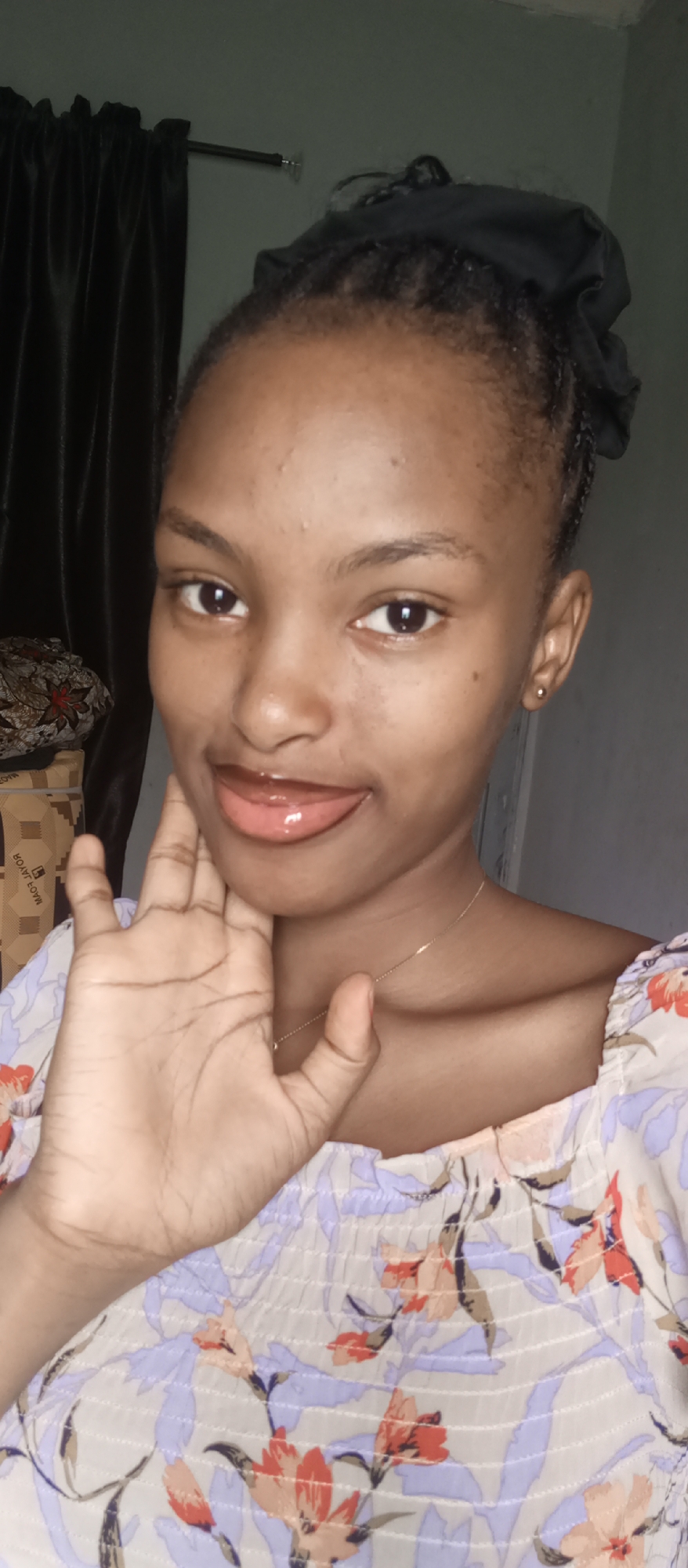 A young woman with dark skin and dark eyes is centered in a close-up, eye-level indoor selfie. She has dark hair styled in braids, pulled back, with a dark scrunchie or fabric visible at the top of her head. Her face shows a light complexion, clear skin with a few minor blemishes, and dark, defined eyebrows. Her lips are glossy, and she has her tongue playfully peeking out. She is wearing a light-colored top with a floral pattern featuring shades of purple, red-orange, and olive green, and a thin gold chain necklace. A small gold stud earring is visible in her right ear. Her left hand is raised to her face, with fingers lightly resting near her jawline.
The background is simple, with a dark curtain hanging from a rod on the left side. A partially visible, light-colored box with some patterned fabric (possibly an African print) on top is seen behind her left shoulder. The word "ROYAL" is visible in inverted text on the box, along with other unreadable text. The wall behind her is a muted, light color, suggesting an indoor setting. The overall lighting is soft and even, without harsh shadows. The woman appears cheerful and relaxed, engaging directly with the camera.
