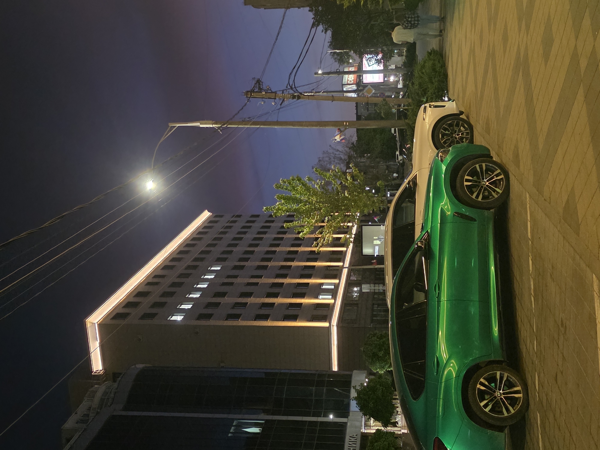 The image shows an urban scene at night. A tall building with illuminated windows and a lit top edge is visible in the background. Two cars are parked on the sidewalk: one green and one white. Streetlights illuminate the scene. Overhead power lines are visible against the night sky. In the background, there are billboards and signs, suggesting a commercial area. Pedestrians are walking on the sidewalk in the distance. The image was taken in the city of Krasnodar, Russia.