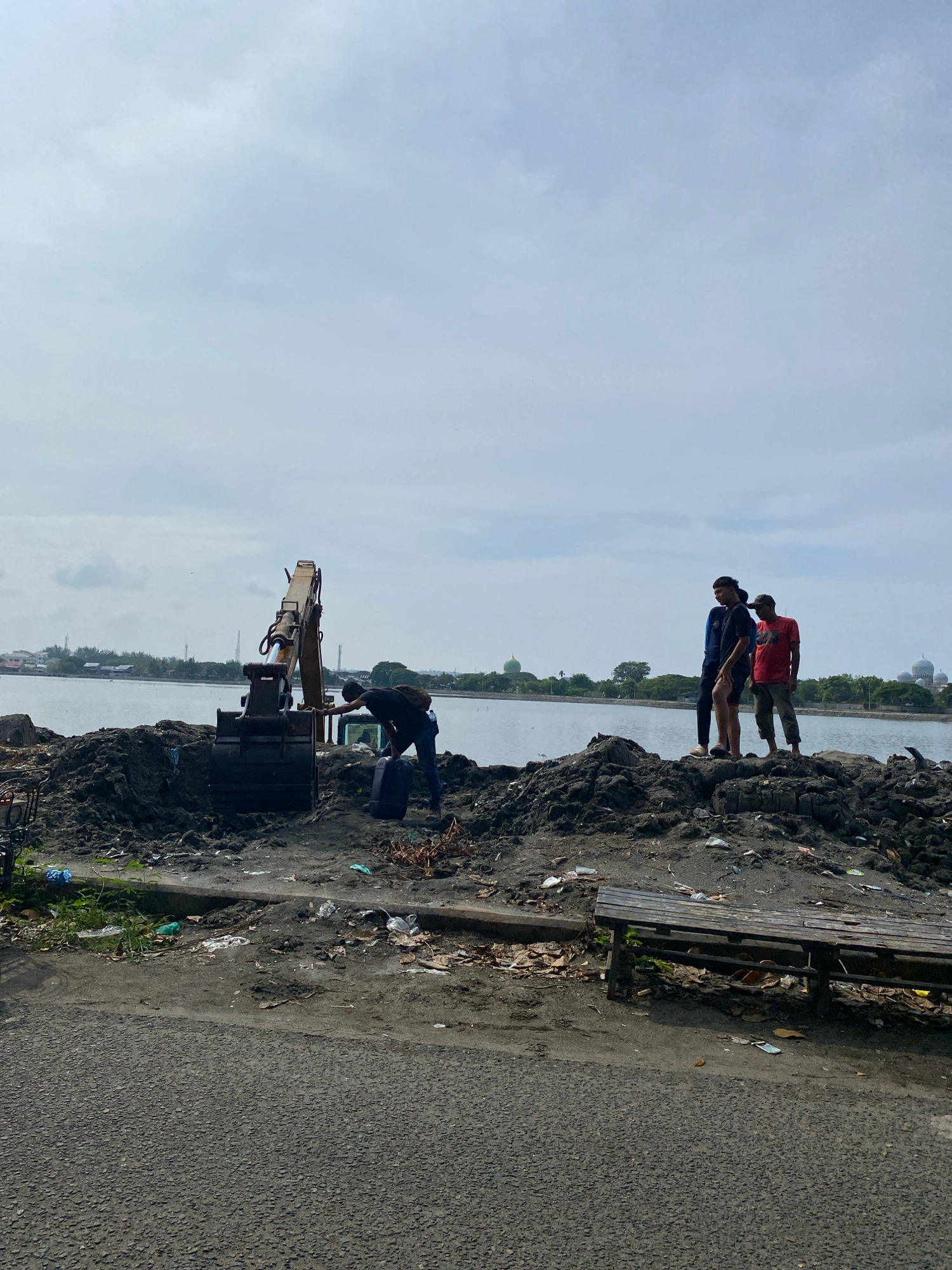The image depicts an outdoor scene near a body of water, likely a river or bay, in Lhokseumawe, Indonesia. The foreground features a paved road, bordered by a dirt area filled with debris, including plastic bags, leaves, and various trash. A wooden bench is situated on the edge of this debris-filled area.
Dominating the mid-ground is a pile of dark soil or mud, with an excavator machine partially embedded in it. A person wearing a black shirt and backpack is bent over, seemingly interacting with something near the excavator's bucket. Further to the right, on top of the soil pile, stand three young men. One is wearing a blue shirt and shorts, another a dark shirt and shorts, and the third a red shirt and dark pants. They are looking out towards the water.
In the background, a calm body of water stretches across the horizon. On the opposite bank, a line of trees and buildings is visible, including several distinct domed structures. The sky is overcast with scattered clouds. The overall impression is one of an industrial or coastal area undergoing some form of excavation or land manipulation, with casual human presence in the vicinity.