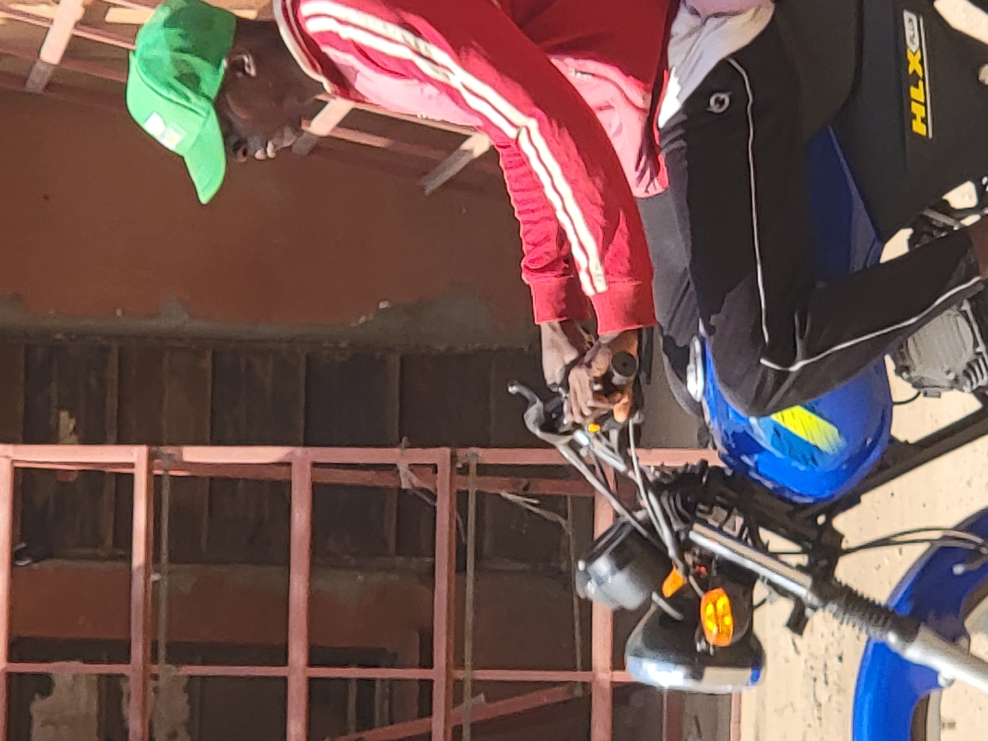 A man on a blue and black motorcycle in Maiburiji, Nigeria. He is wearing a red tracksuit with white stripes and a green baseball cap. He is holding the handlebars of the motorcycle. The background shows a building with a red structure.