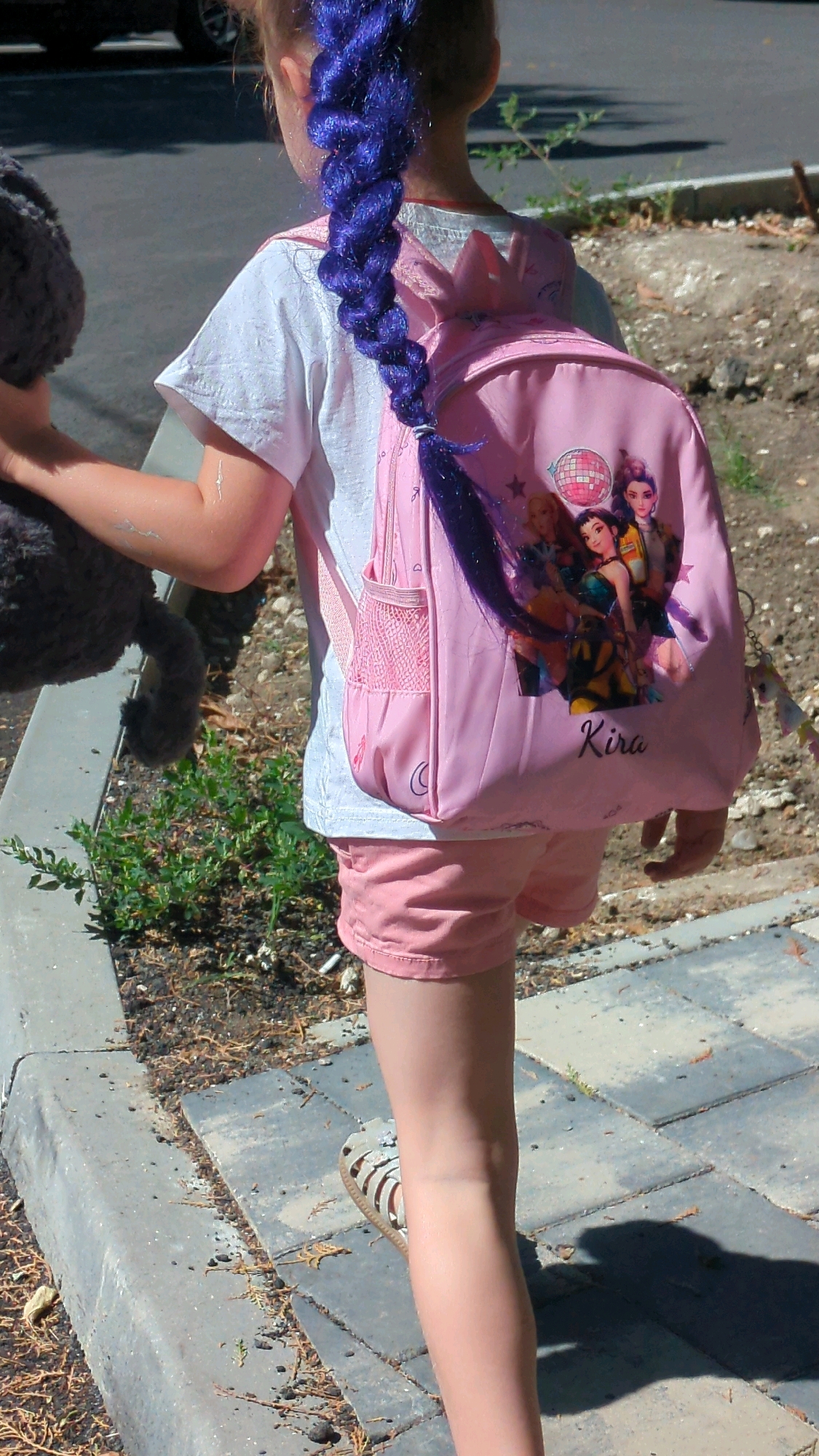 A young girl is seen from behind, walking away from the camera on a sidewalk. She has bright purple hair styled in a long braid that hangs down her back. She is wearing a white t-shirt, pink shorts, and sandals. A pink backpack with images of cartoon characters and the name "Kira" is on her back. She is also carrying a gray stuffed animal in her left arm. The environment appears to be an urban setting, with a concrete curb and paving stones visible, as well as some sparse vegetation.