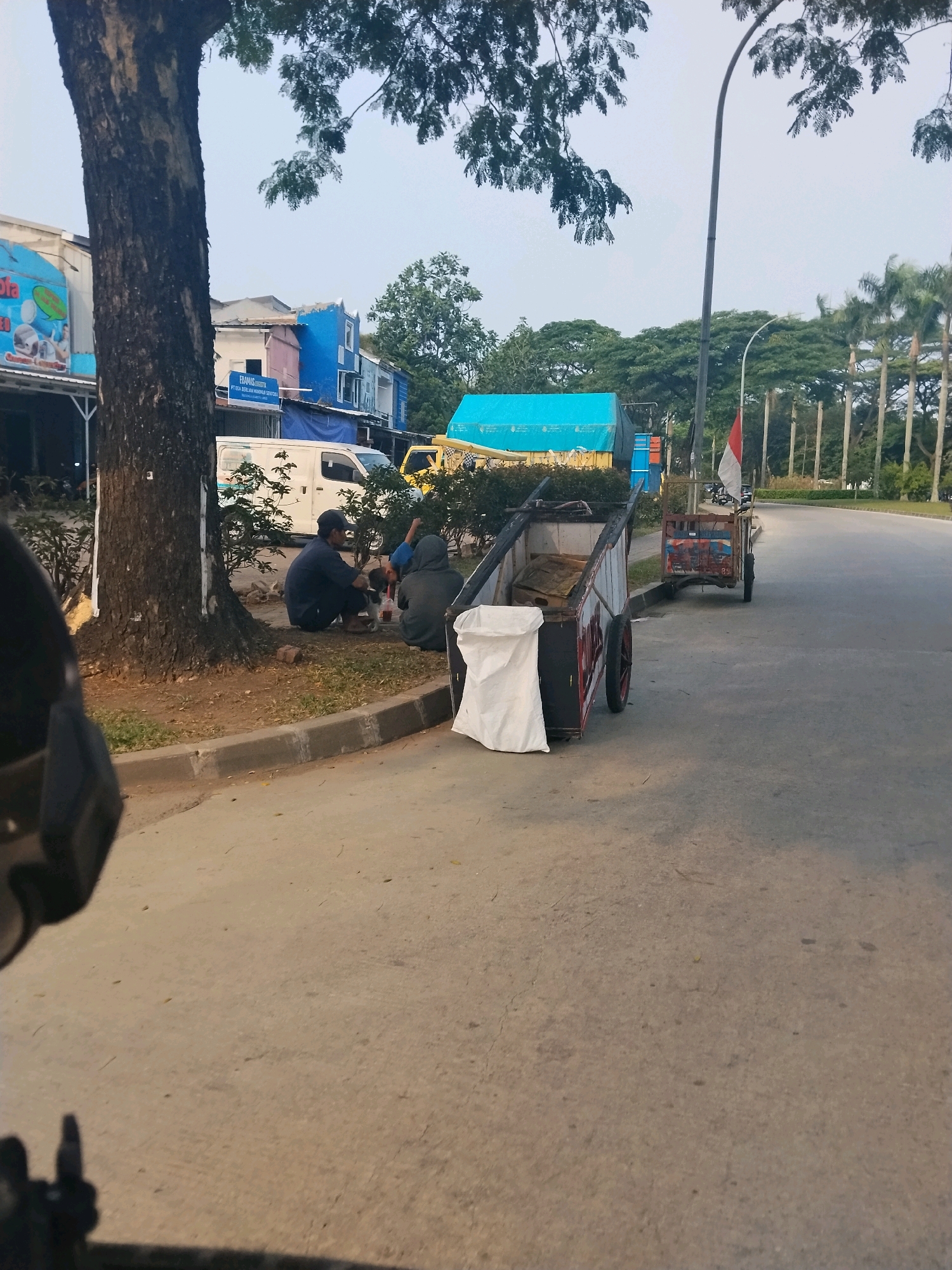 Two individuals are seated on the ground near the base of a large tree by the side of a paved road. One person is wearing a dark blue shirt and a dark cap, and the other is wearing a grey hooded jacket. They appear to be interacting with a small object or animal on the ground between them. In the foreground, a rustic cart with red wheels and a white bag is parked on the road. Behind the cart, another similar cart is visible, decorated with a red and white flag. Several vehicles, including a white van and a yellow truck with a blue tarp, are parked further back along the roadside, near a row of buildings. Palm trees and other lush foliage line the far side of the road, suggesting a tropical or subtropical urban environment. The sky is overcast, casting a diffused light on the scene. The presence of the Indonesian flag on one of the carts indicates a location within Indonesia. Given the context provided, the scene is in Cikupa, Indonesia.