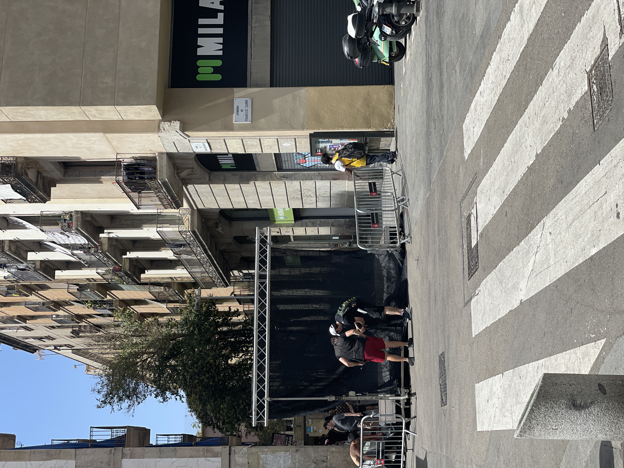 In Barcelona, Spain, on a sunny day, people are gathered around a temporary stage set up on a street. The stage has a large black backdrop supported by a metal truss structure. Two men, one wearing red shorts and a white cap, and another in a black vest and shorts, are interacting near the stage. Other individuals are visible in the background, some observing the setup and others moving along the street.
The scene is located in a city environment with multi-story buildings lining the street. One building features several balconies with railings, and some laundry is visible on one of the balconies. A store with a black facade and a prominent green logo that reads "MILAN" is situated on the corner of the building. A street sign on the corner reads "AVINGUDA DE FRANCESC CAMBO".
In the foreground, a pedestrian crosswalk with white stripes is painted on the asphalt. Several metal barriers are erected on the sidewalk and street, likely for crowd control or to delineate the event area. A gray concrete bollard is positioned near the crosswalk. Two motorcycles, one light green and the other dark, are parked on the side of the street. The ground is paved with asphalt, and manhole covers are visible. Trees with green foliage are present, adding to the urban landscape. The sky is clear and blue.