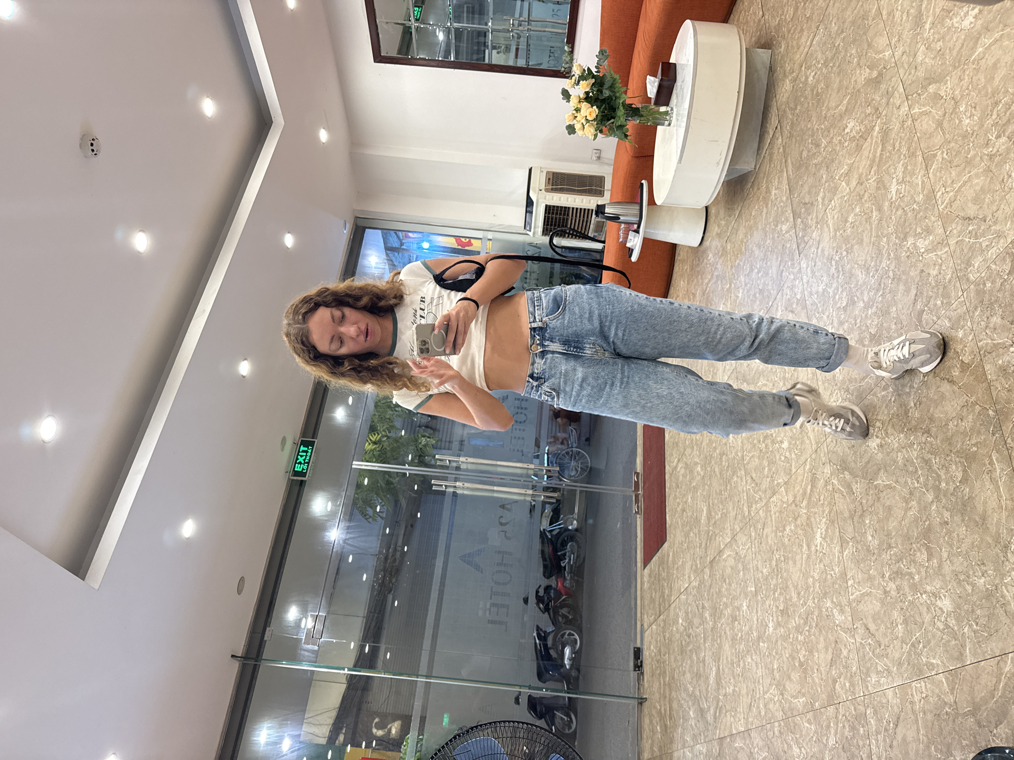 A young woman with curly brown hair stands in a modern hotel lobby, looking down at her phone. She is wearing a white crop top with a green trim and blue denim jeans. Her left hand is raised, holding her phone, while her right hand is resting on the phone. She is wearing white sneakers with grey accents and white socks. Behind her, a glass door reveals a street scene with motorcycles parked outside. The lobby features beige marble flooring, a white ceiling with recessed lighting, and an orange sofa with white side tables in the background. A vase of yellow roses sits on one of the tables. The "EXIT" sign is visible above the glass door. The hotel appears to be located in Hanoi, Vietnam, as indicated by the Vietnamese text on the exit sign.