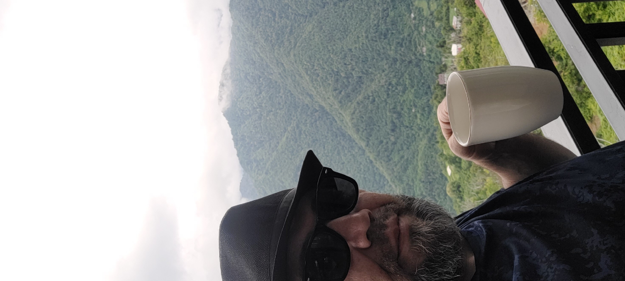 A man wearing a black hat and sunglasses is holding a white mug, with a misty, green mountainous landscape in the background. The scene appears to be from a balcony or outdoor seating area, with a railing in the foreground. The man has a beard and is wearing a dark blue t-shirt. The mountains are covered in lush vegetation, and the sky is overcast with clouds. The location is identified as Milisi, Georgia.