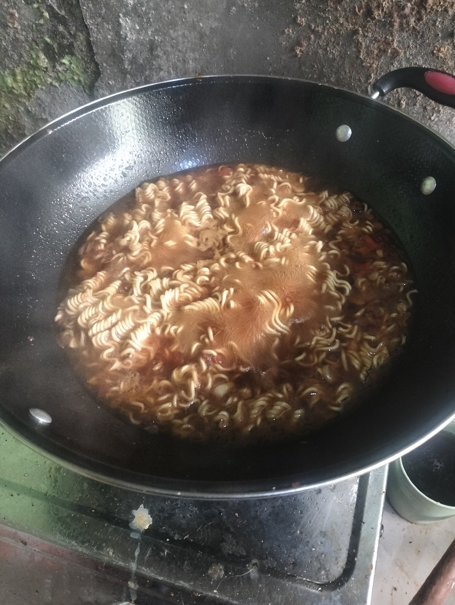 In Jetis, Indonesia, a close-up shot captures a black frying pan filled with instant noodles and broth, actively simmering on a stovetop. The noodles, characteristic of instant ramen, are coiled and submerged in a reddish-brown liquid, with visible steam rising, indicating they are being cooked. The pan has silver rivets securing its handle, which is partially visible on the right side of the frame and appears to have a red grip. The stovetop beneath the pan is metal and shows signs of use and grime. In the background, a rough, textured surface, possibly a wall or rock formation, is visible, with some patches of green moss or algae. To the right of the pan, a small, light green cylindrical object, likely a cup or container, is present. The overall scene suggests a simple, home-cooked meal in progress.